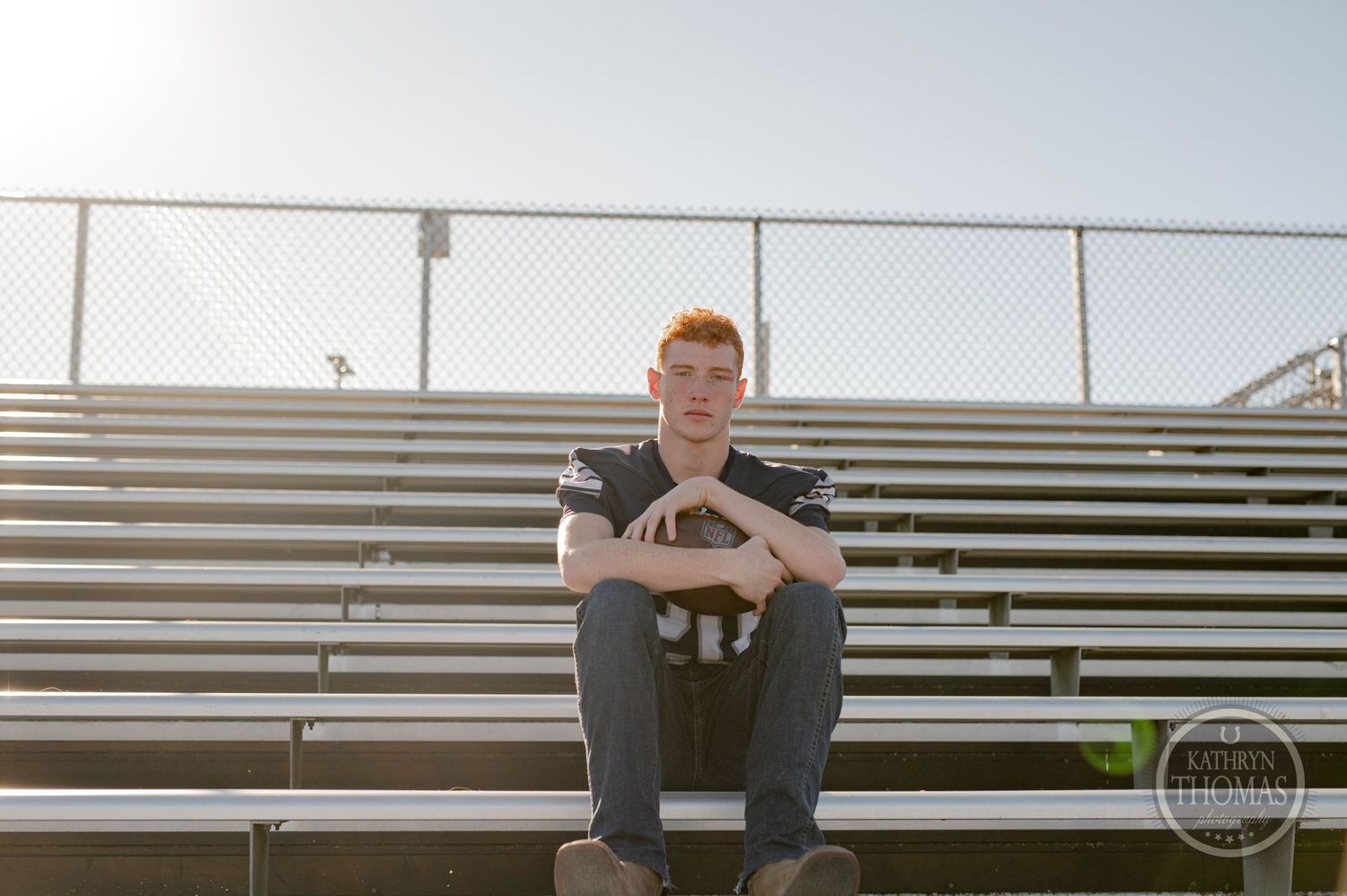 An athlete in a football jersey sits on metal bleachers at a sports field.