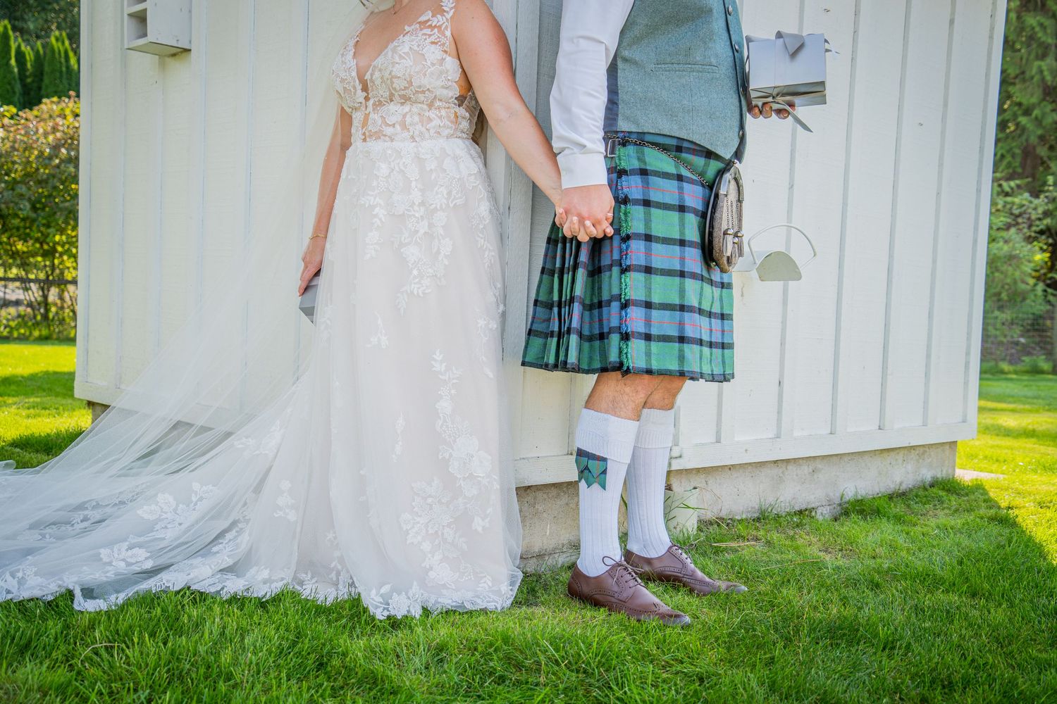 A couple stands together showing a traditional Scottish kilt and flowing white wedding dress train on green grass.