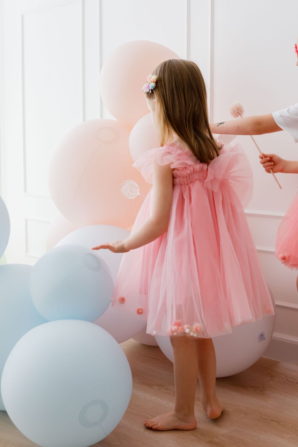A young child in a pink tulle dress stands barefoot among pink and blue balloons in a bright room.