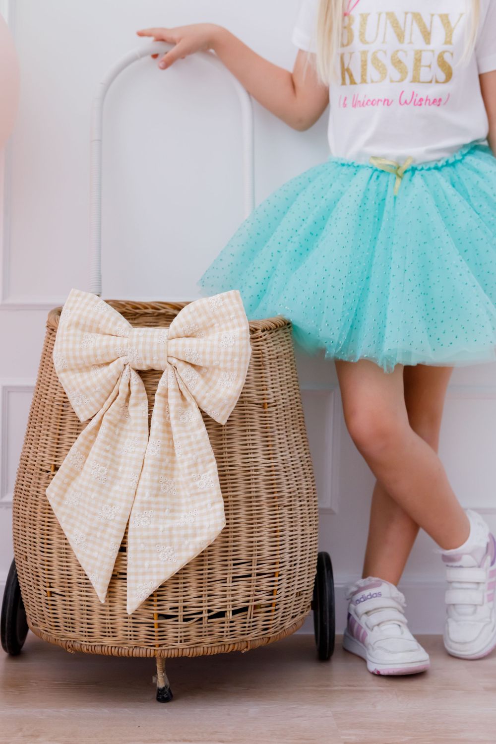 Wicker storage basket with cream bow beside teal tulle skirt and white sneakers on wooden floor.