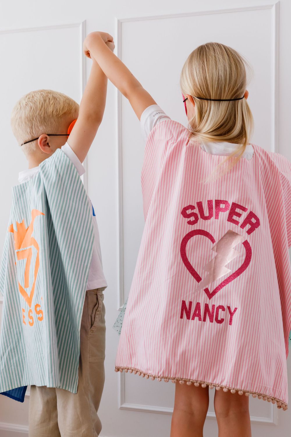 Two children wearing stripe patterned superhero capes in pink and blue colors with text designs standing against a white wall.