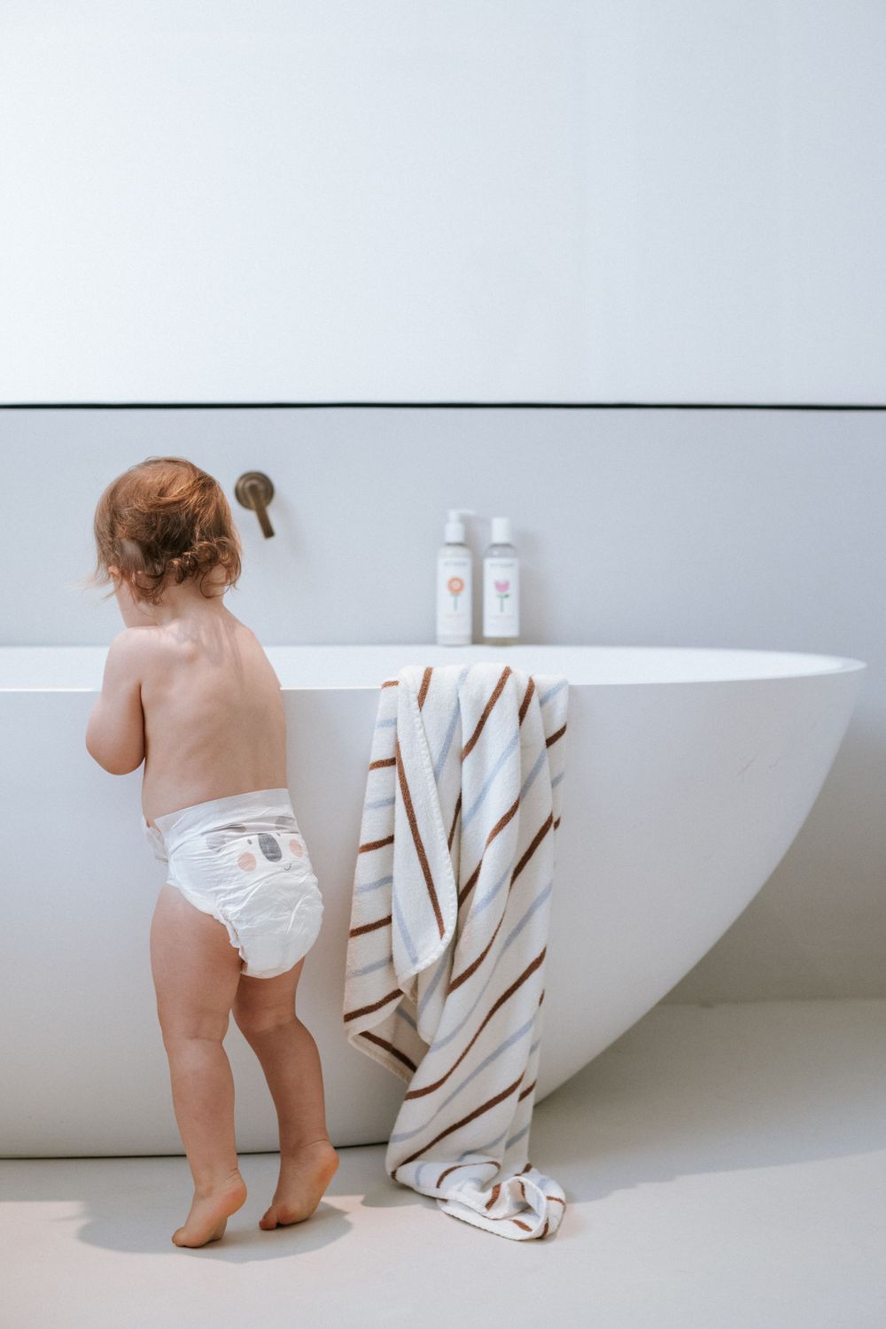 A toddler in a diaper reaches toward a modern white freestanding bathtub with striped towel hanging on the side.