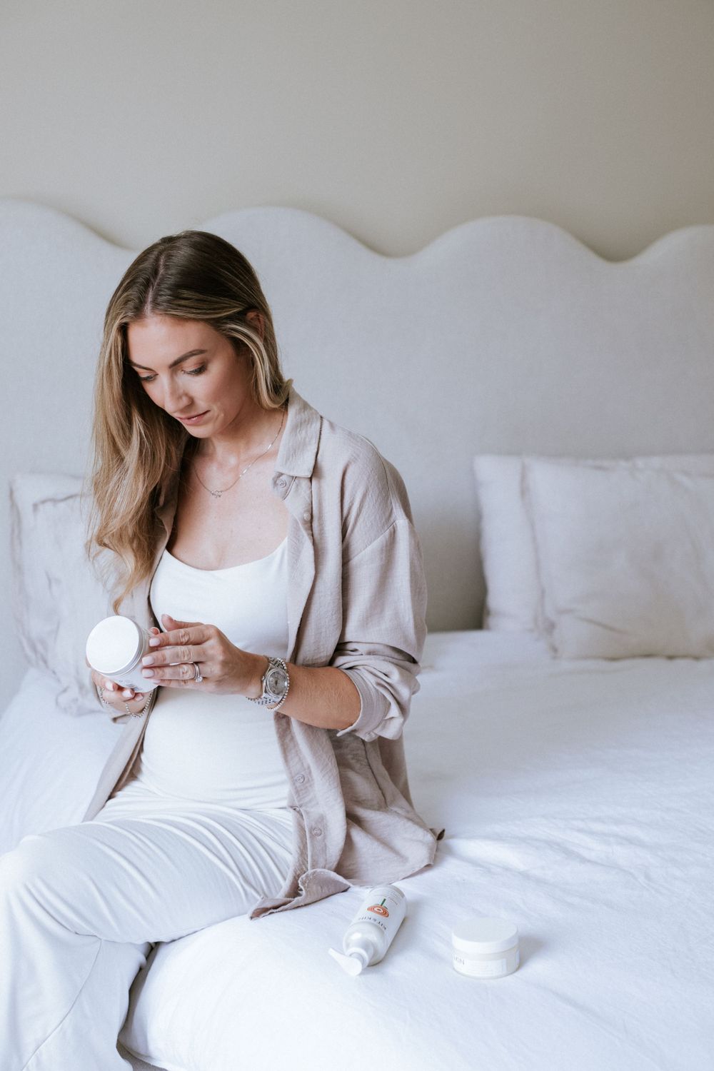 Woman in beige cardigan and white outfit relaxes on white bedding while holding skincare products.