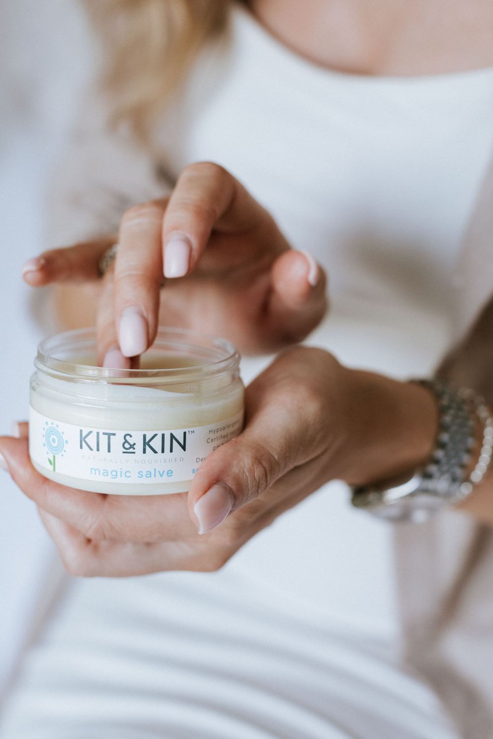 Hands holding a jar of Kit & Kin organic skin salve product while wearing a white garment and bracelet.