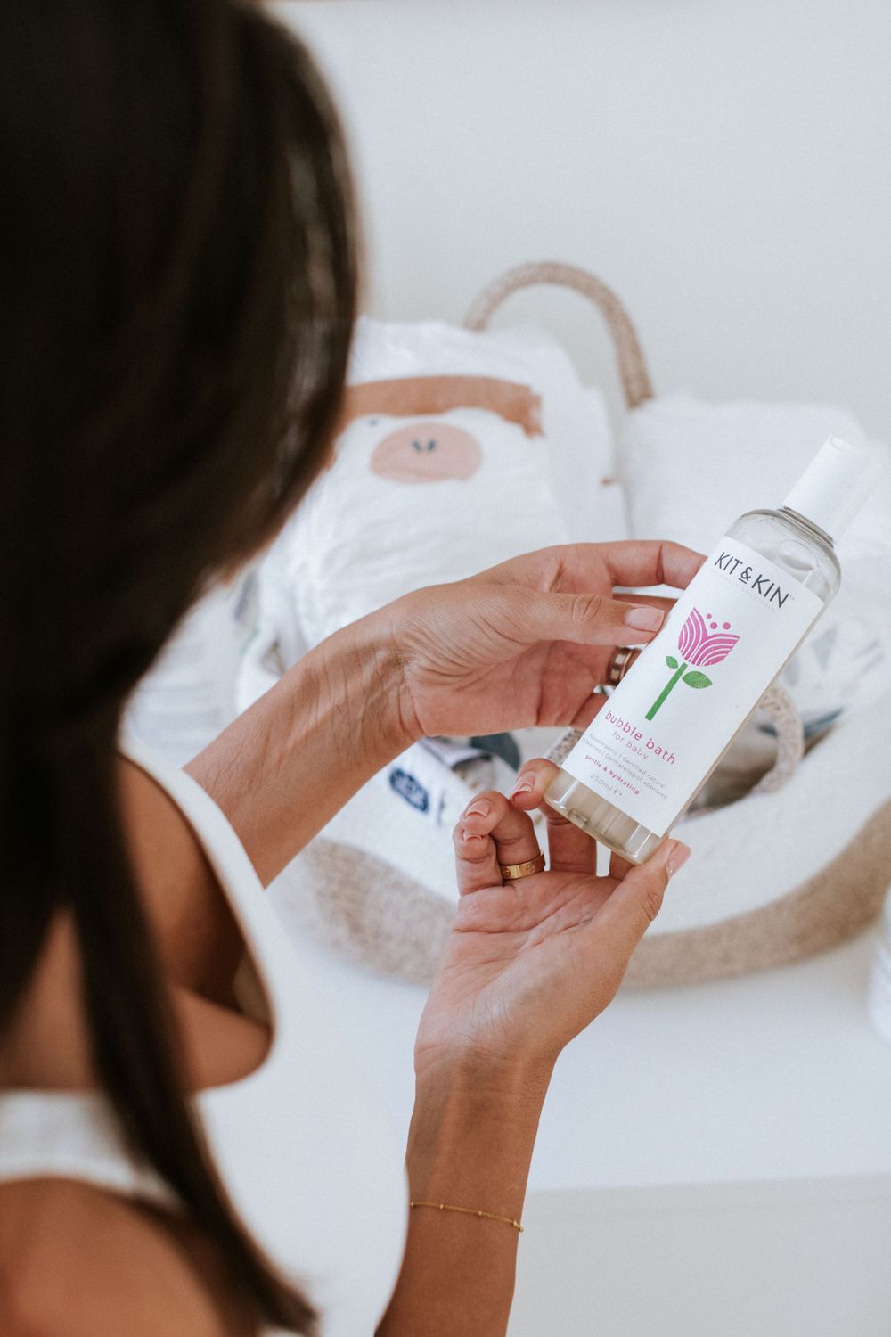 Hands holding a skincare product bottle against a white bathtub background with soft natural lighting.