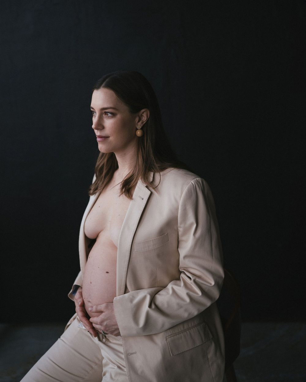 A dramatic maternity portrait against a dark background in beige business attire.