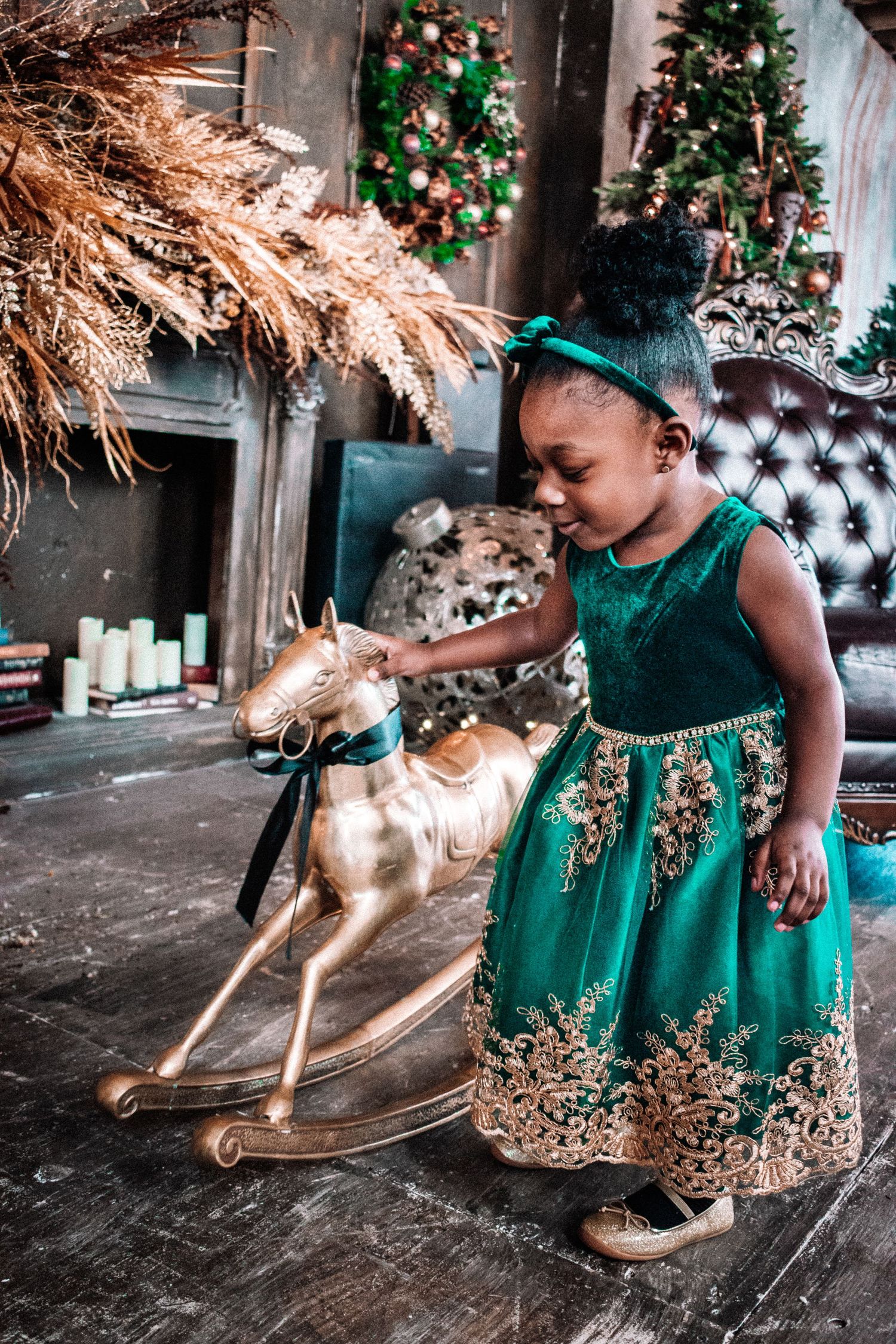 A child in an emerald green and gold holiday dress stands beside a decorative rocking horse with Christmas trees in background. Tacoma photo