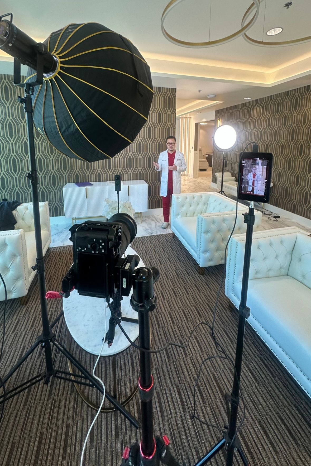 Professional photo studio setup with lighting equipment, white tufted chairs and patterned wallpaper in an elegant room.