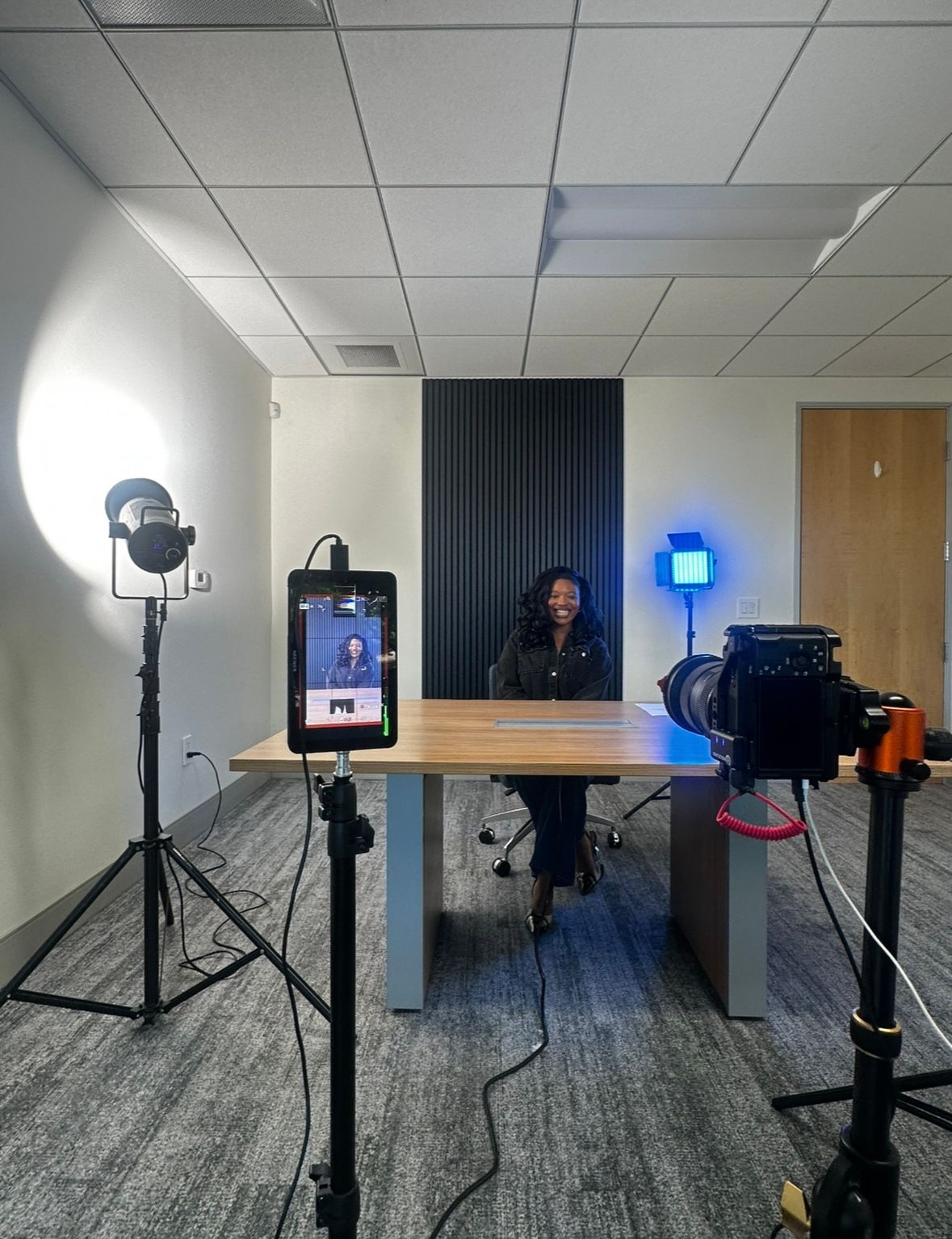 Professional video recording setup in office with lighting equipment, camera gear, and desk against dark accent wall.