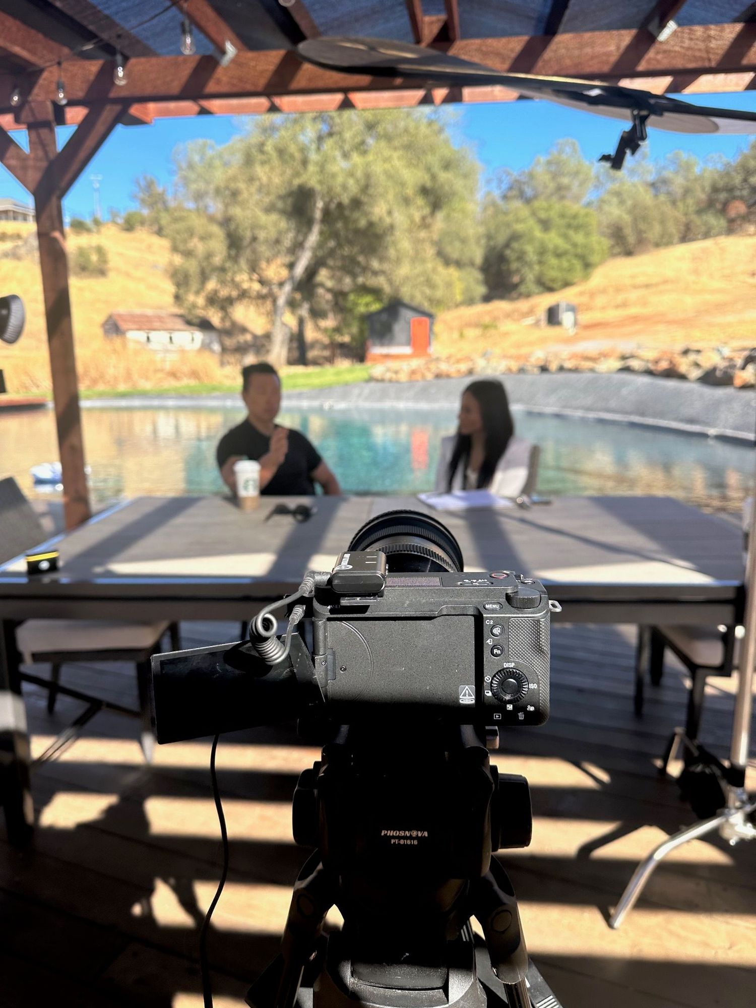 Professional camera setup on tripod filming interview subjects at outdoor poolside location under wooden pergola.