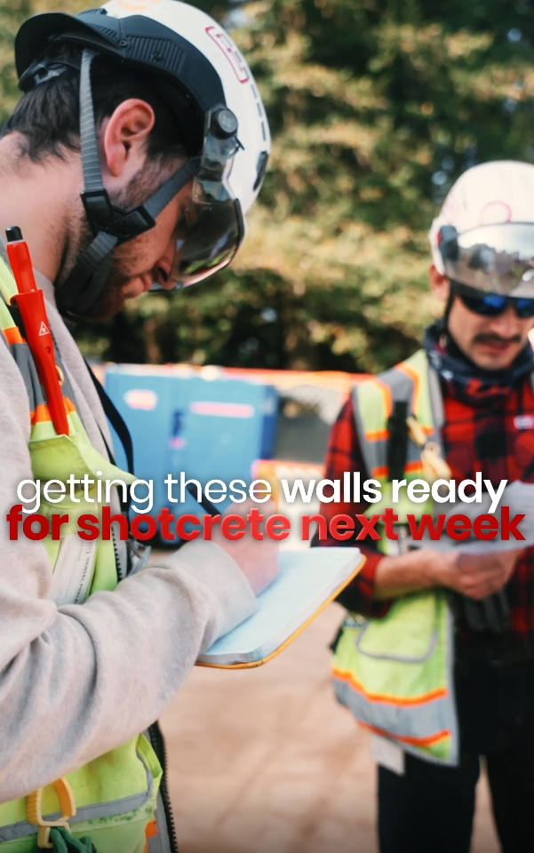 Two construction workers in safety gear review blueprints on a job site, preparing walls for shotcrete application.