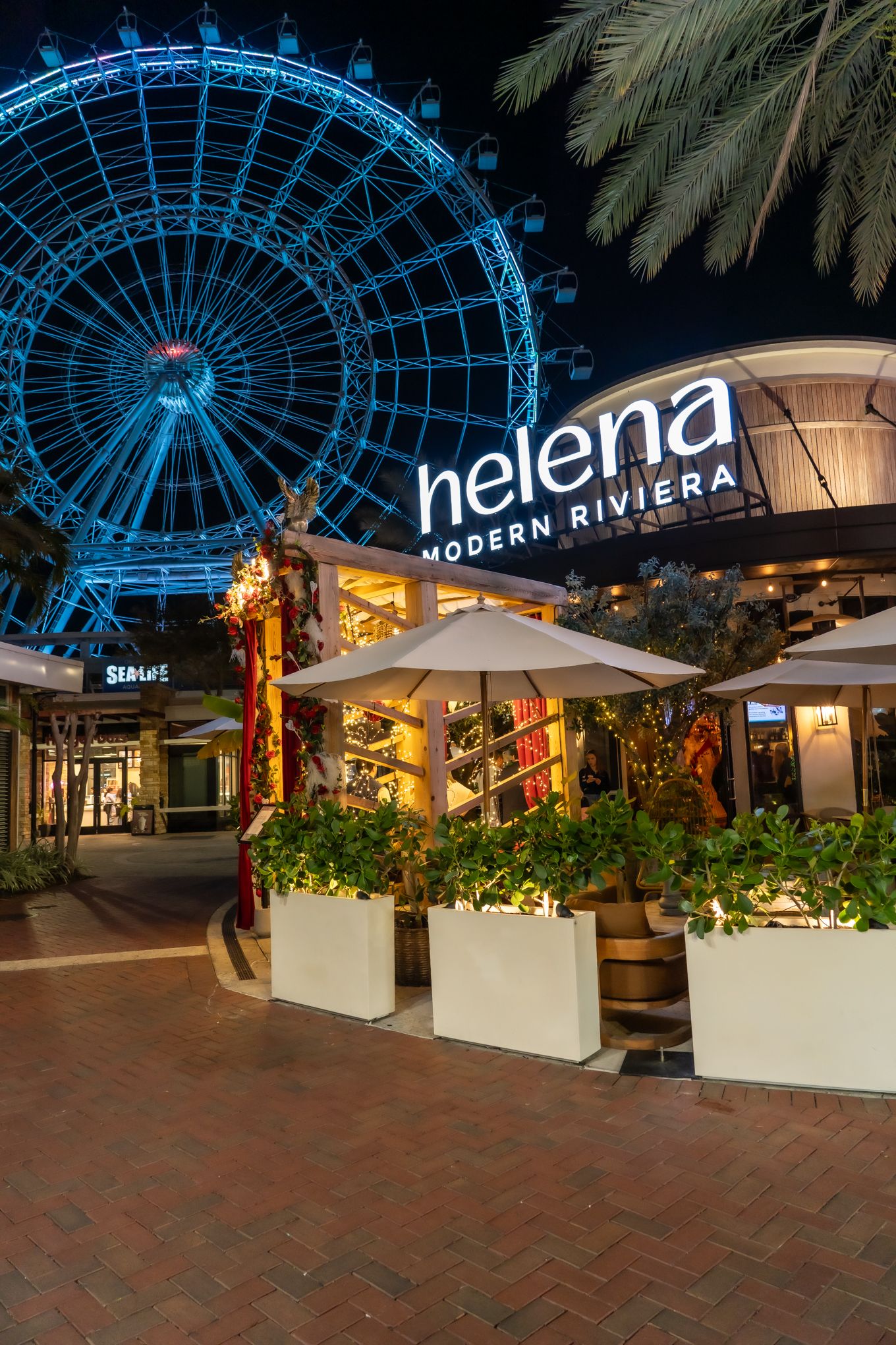 Night view of Helena Modern Riviera with illuminated blue Ferris wheel and outdoor dining area.