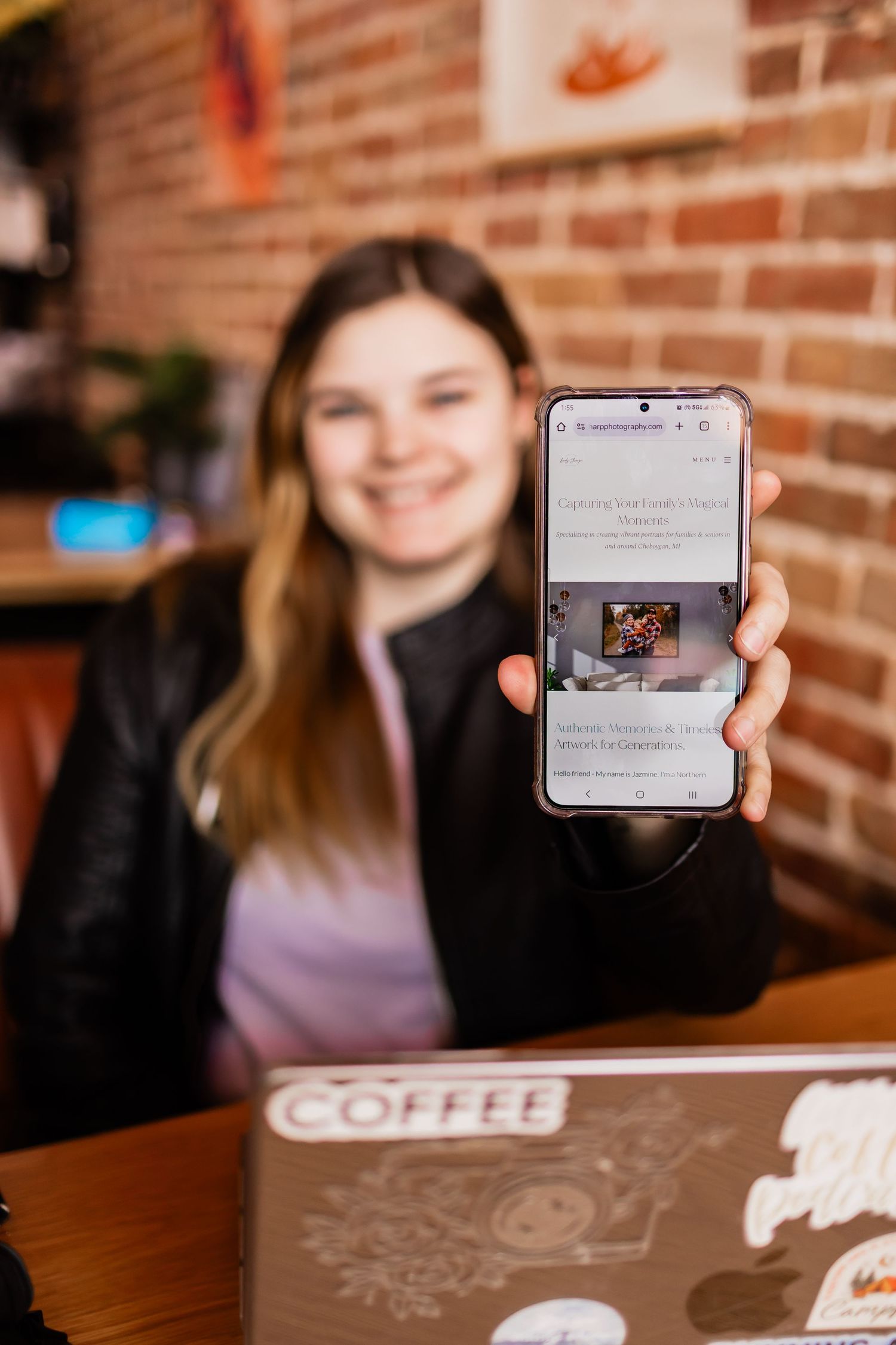 Someone holds up a smartphone displaying content while sitting at a coffee shop table with a brick wall backdrop.