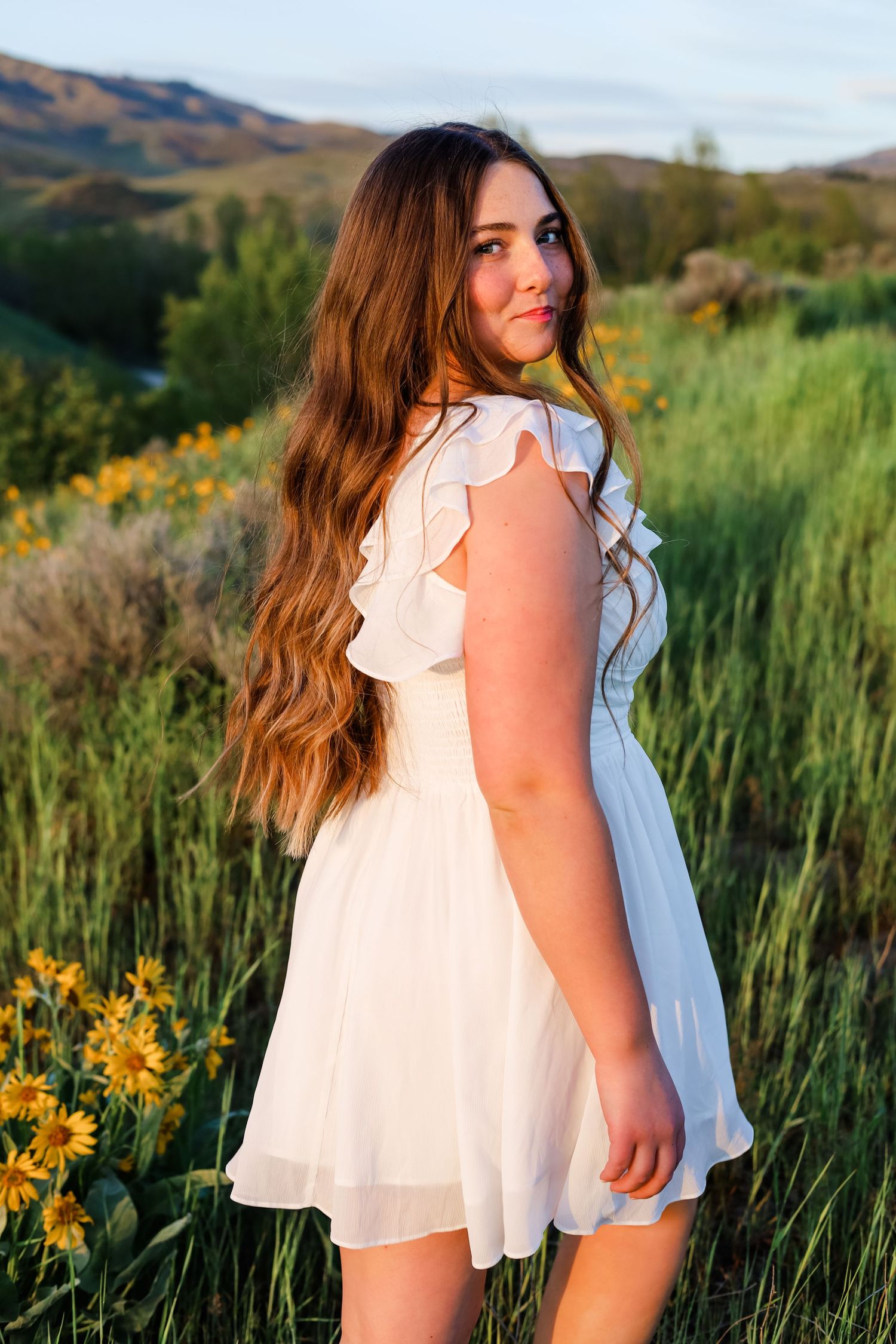 Woman in white flowing dress poses among yellow wildflowers at sunset.