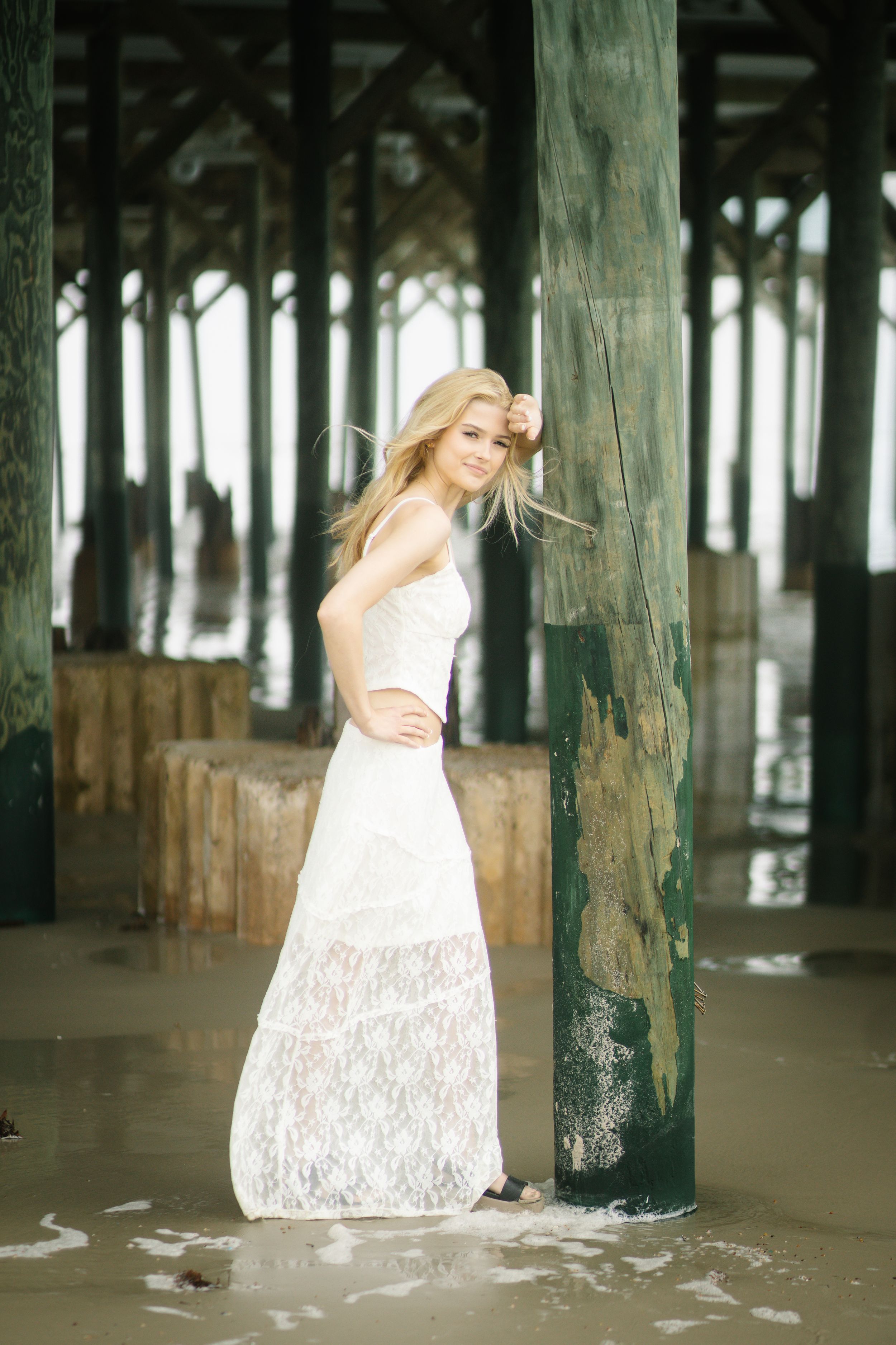 Woman in white lace dress poses under wooden pier pilings with ocean waves washing around her feet.