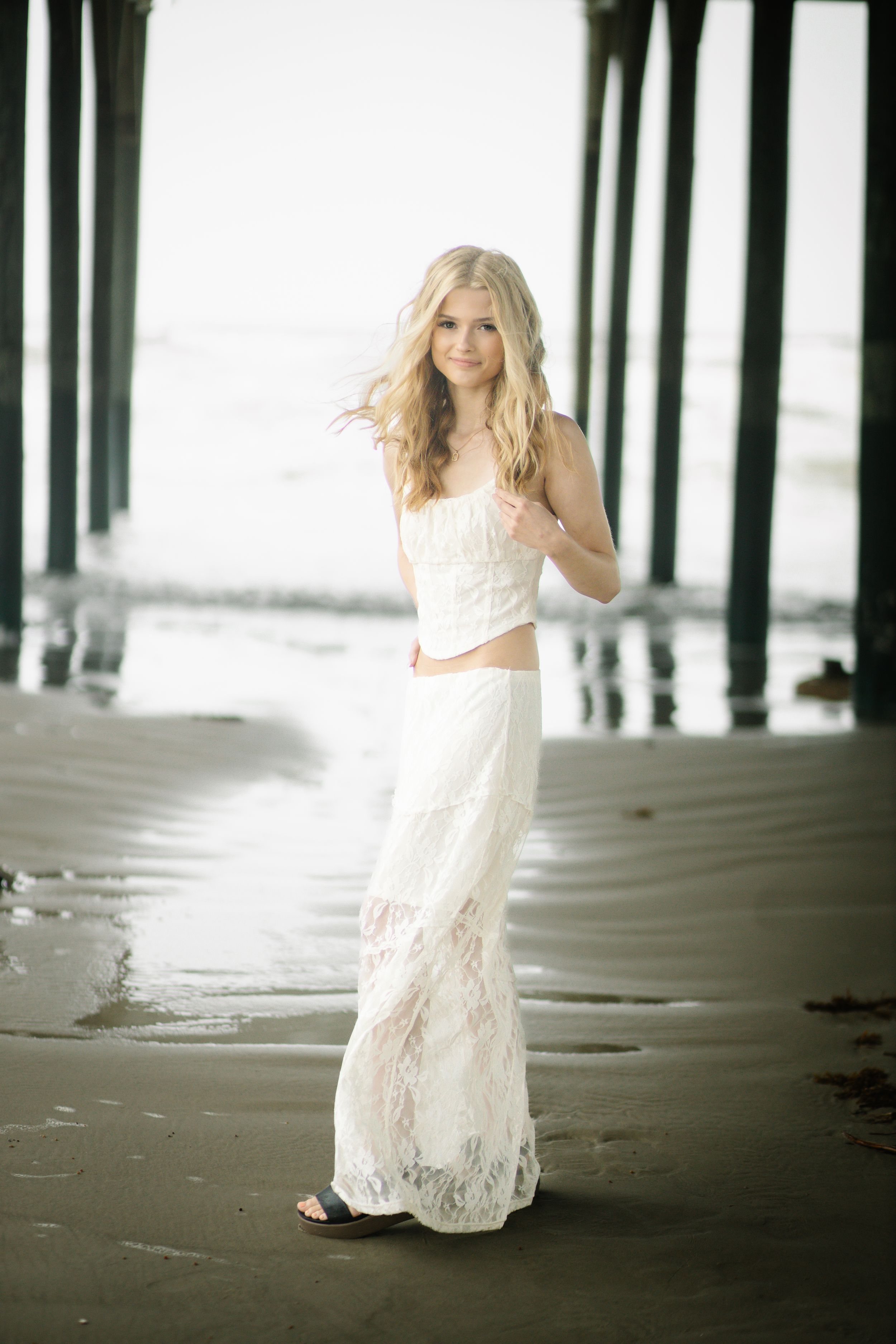 Woman in flowing white lace dress poses beneath pier pillars on sandy beach at sunset.