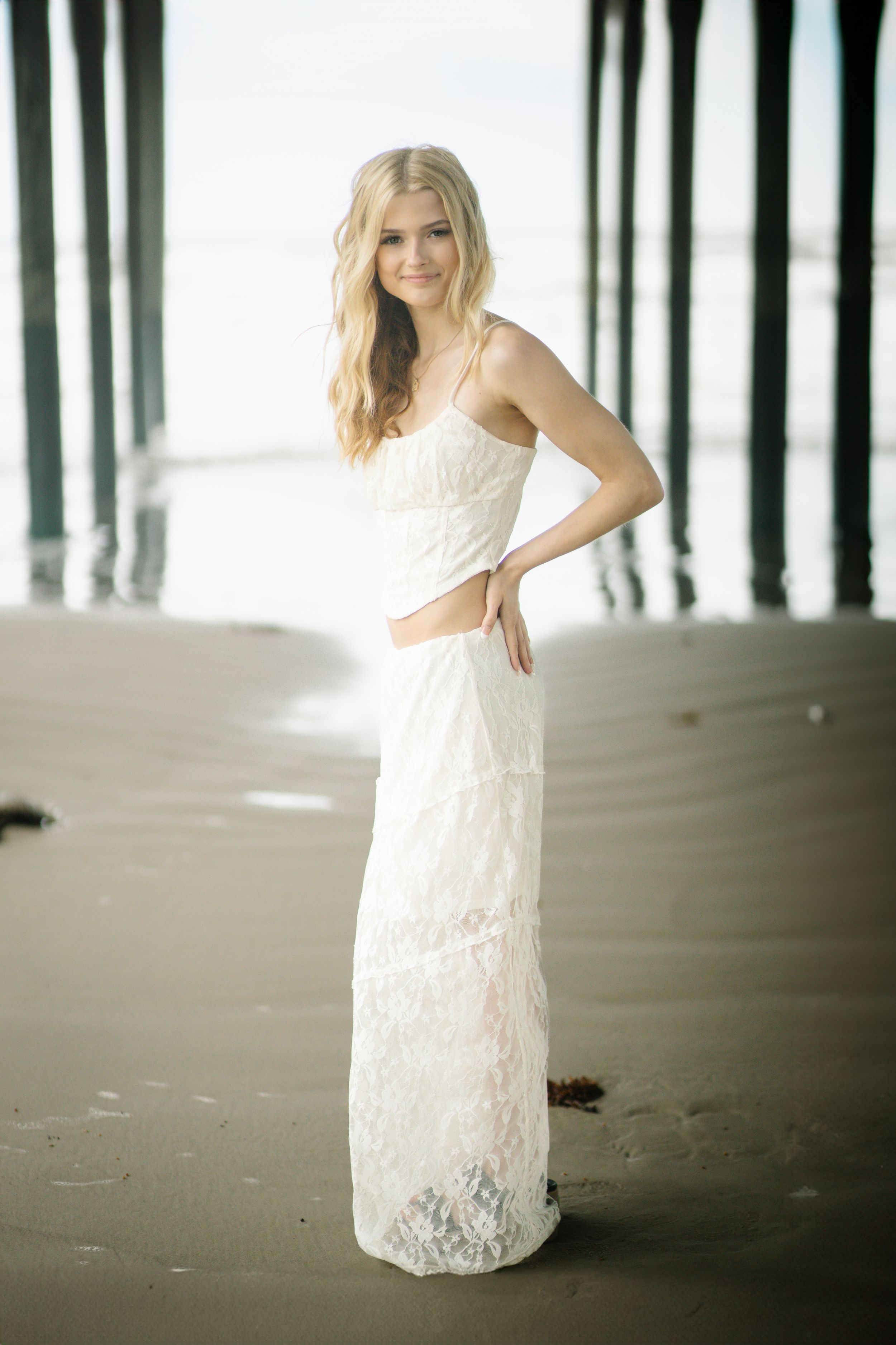 A white lace wedding dress photographed on a beach with dark wooden pier pilings in the background.