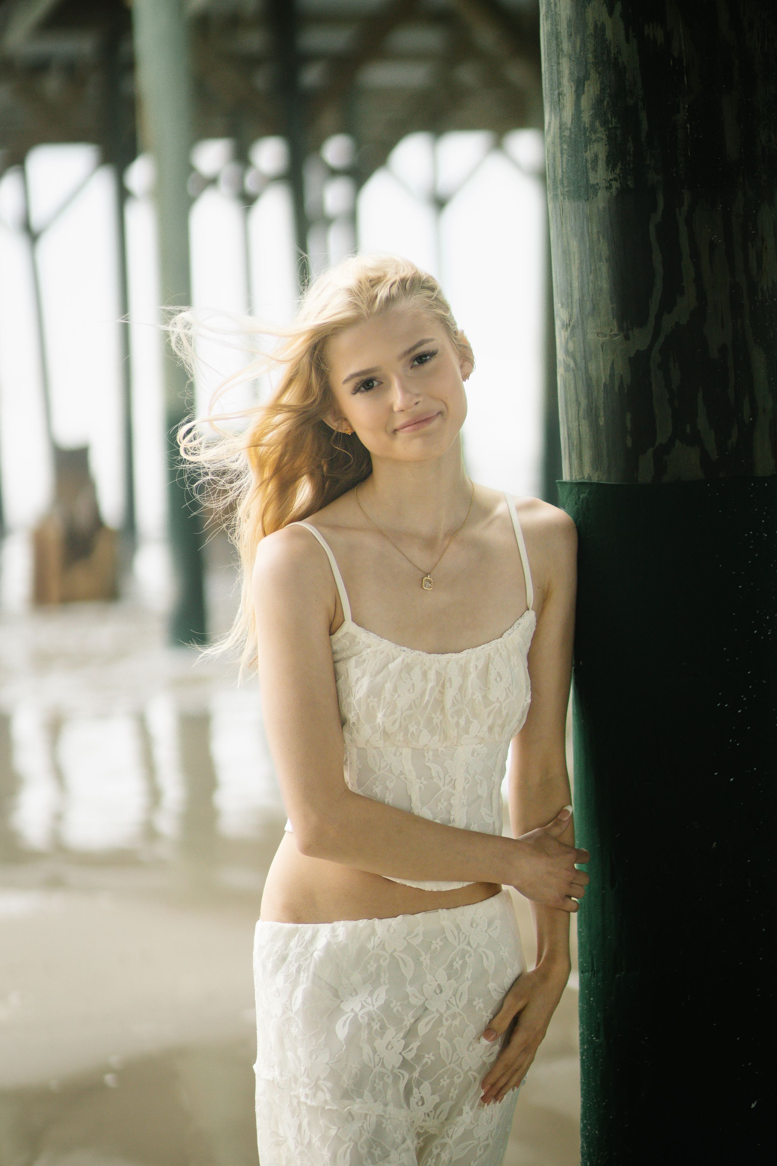 Person in white lace dress poses against wooden pier pillars as wind blows their hair.