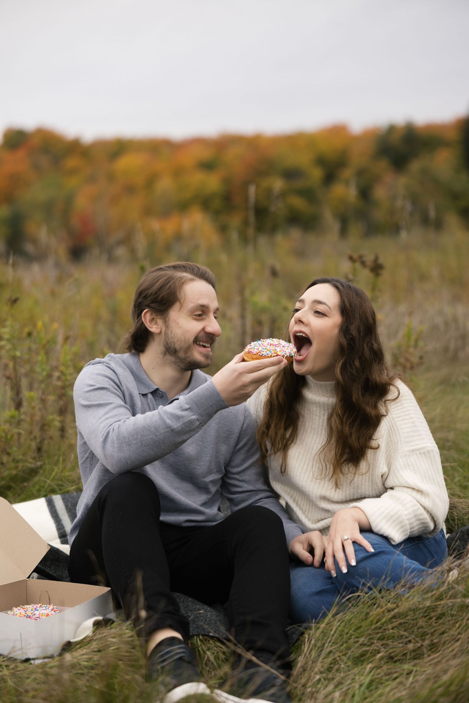 Two people wearing pumpkin masks and fall sweaters pose playfully in an autumn field with colorful foliage in background.