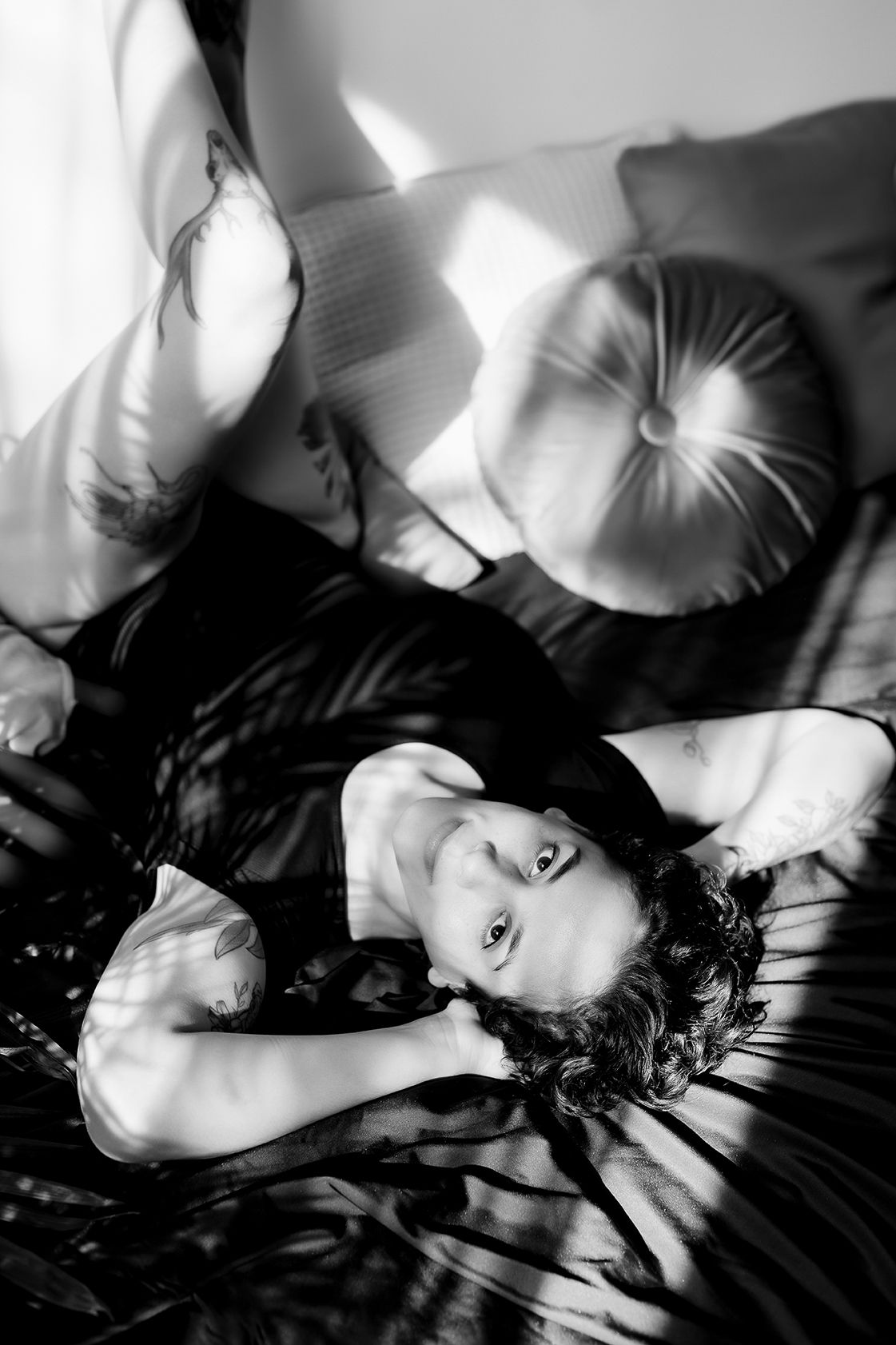 Black and white boudoir photograph with dramatic lighting showing figure reclining on pillows in intimate setting.