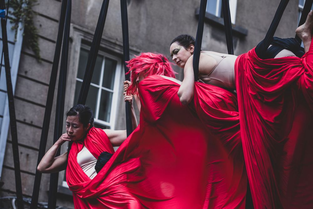 Performers in flowing red fabric costumes dance and move dramatically against an urban building backdrop.