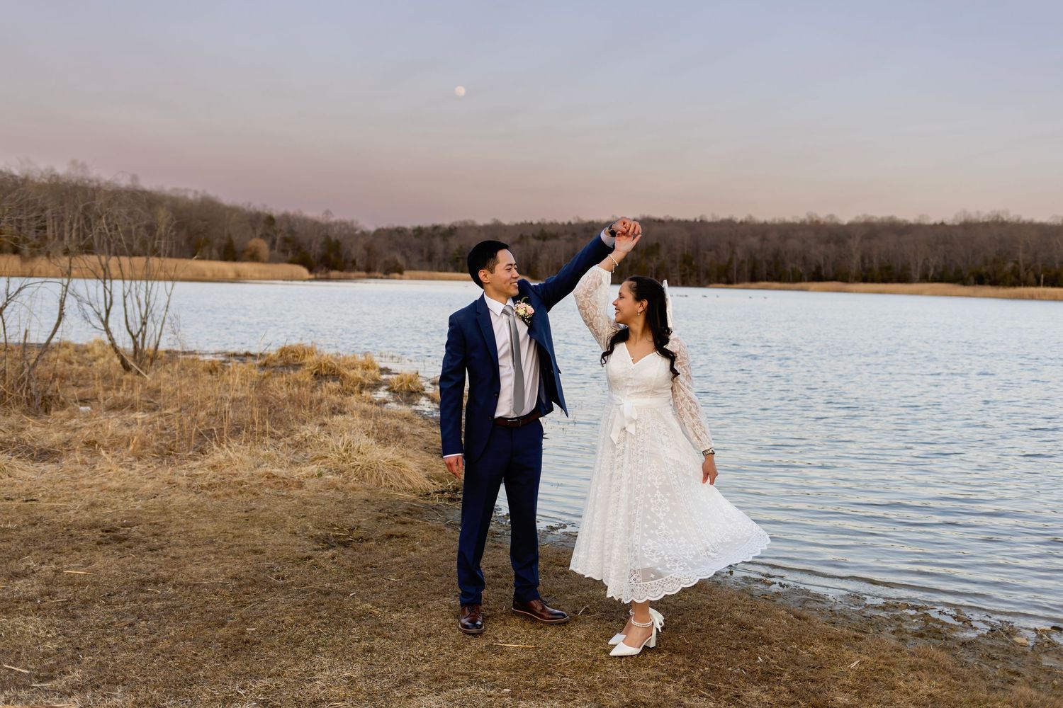 A couple in formal wedding attire dances joyfully by a scenic lake at sunset with bare winter trees in the background.
