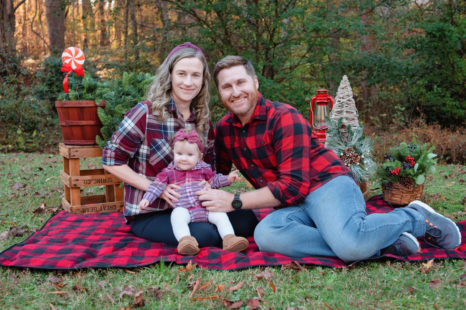 Family wearing red plaid flannel sits on picnic blanket with holiday decorations in autumn forest setting.