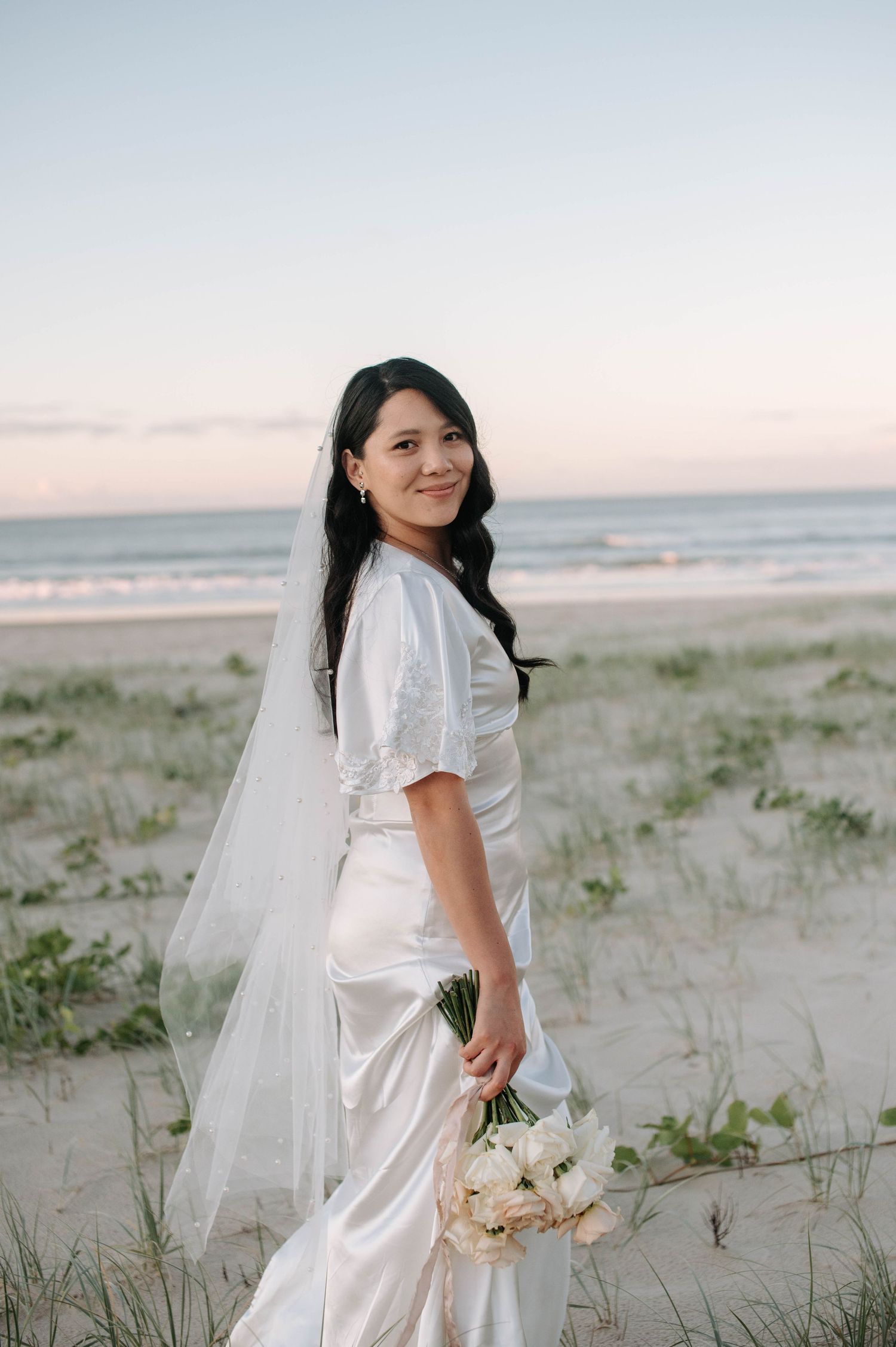 A bride wearing a white satin dress and veil holds a bouquet of white flowers on a serene beach at sunset.