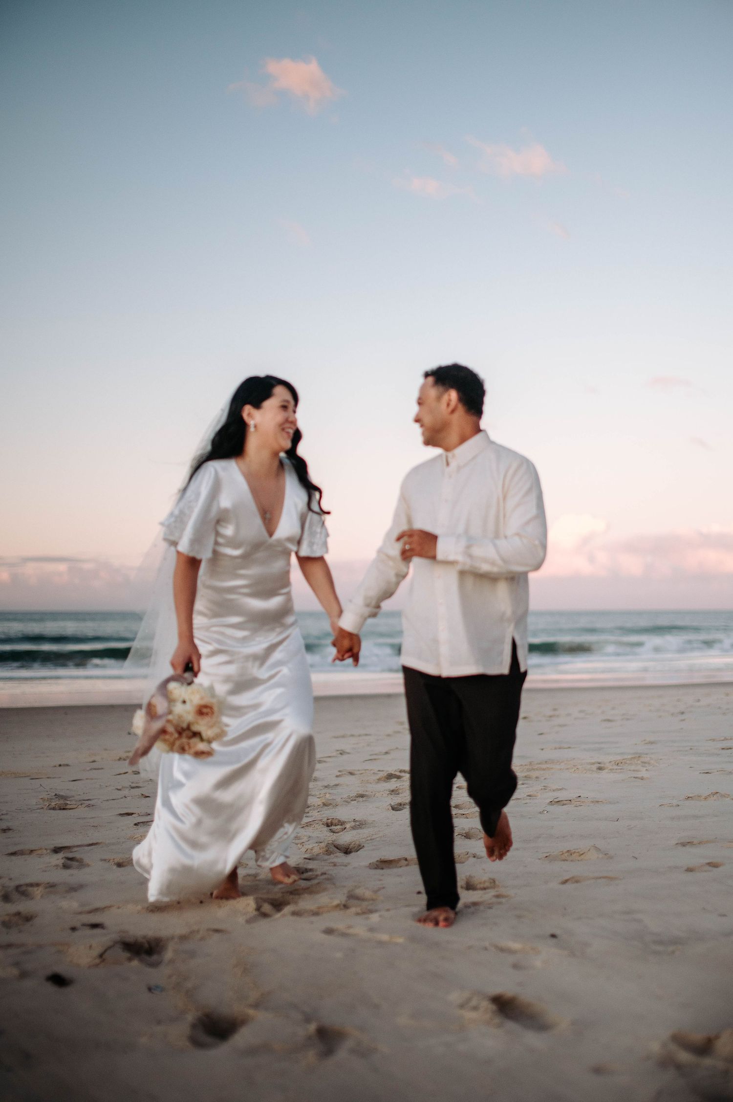 A couple runs joyfully along the beach at sunset, with the bride in a flowing white dress holding a bouquet.