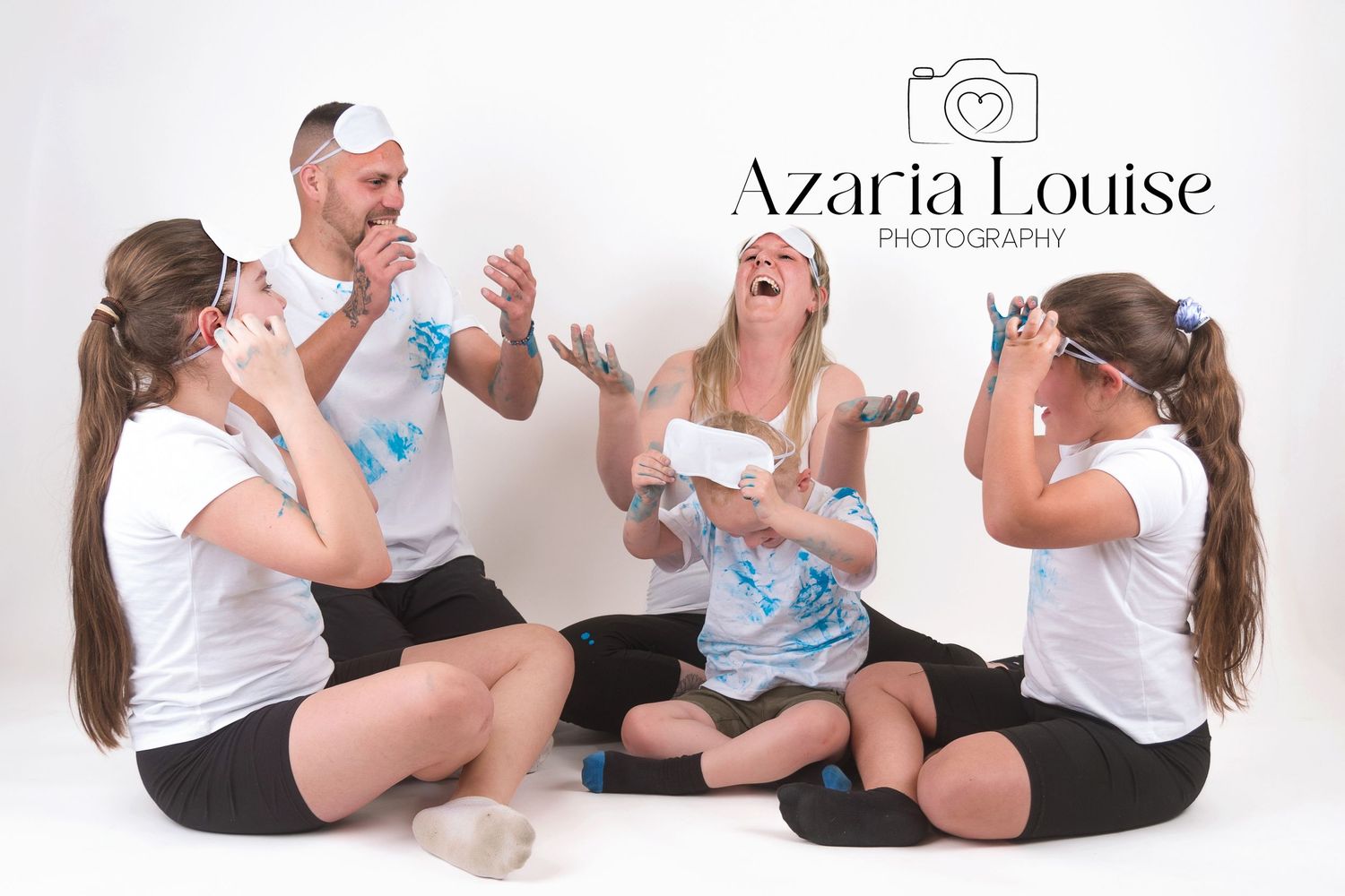 A group of people in white t-shirts sit on the floor laughing and playing with baby powder in a studio setting.