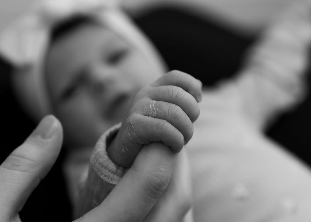 Black and white photo of newborn baby grasping adult's finger, showing tiny hand and wrist bracelet.