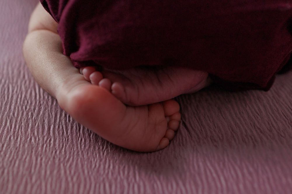 Close up of tiny baby feet on pink textured bedding with burgundy fabric draped across.