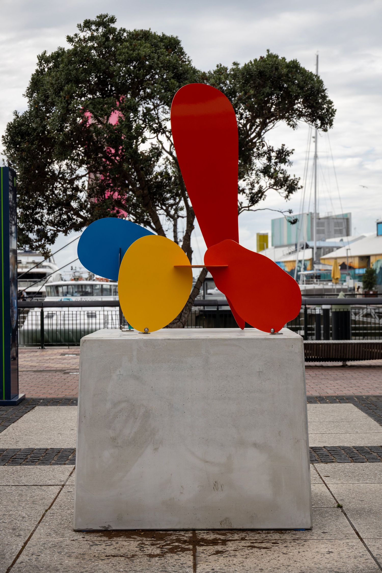 Abstract sculpture with red, yellow, and blue shapes mounted on a white block in a harbourside plaza with trees and buildings.