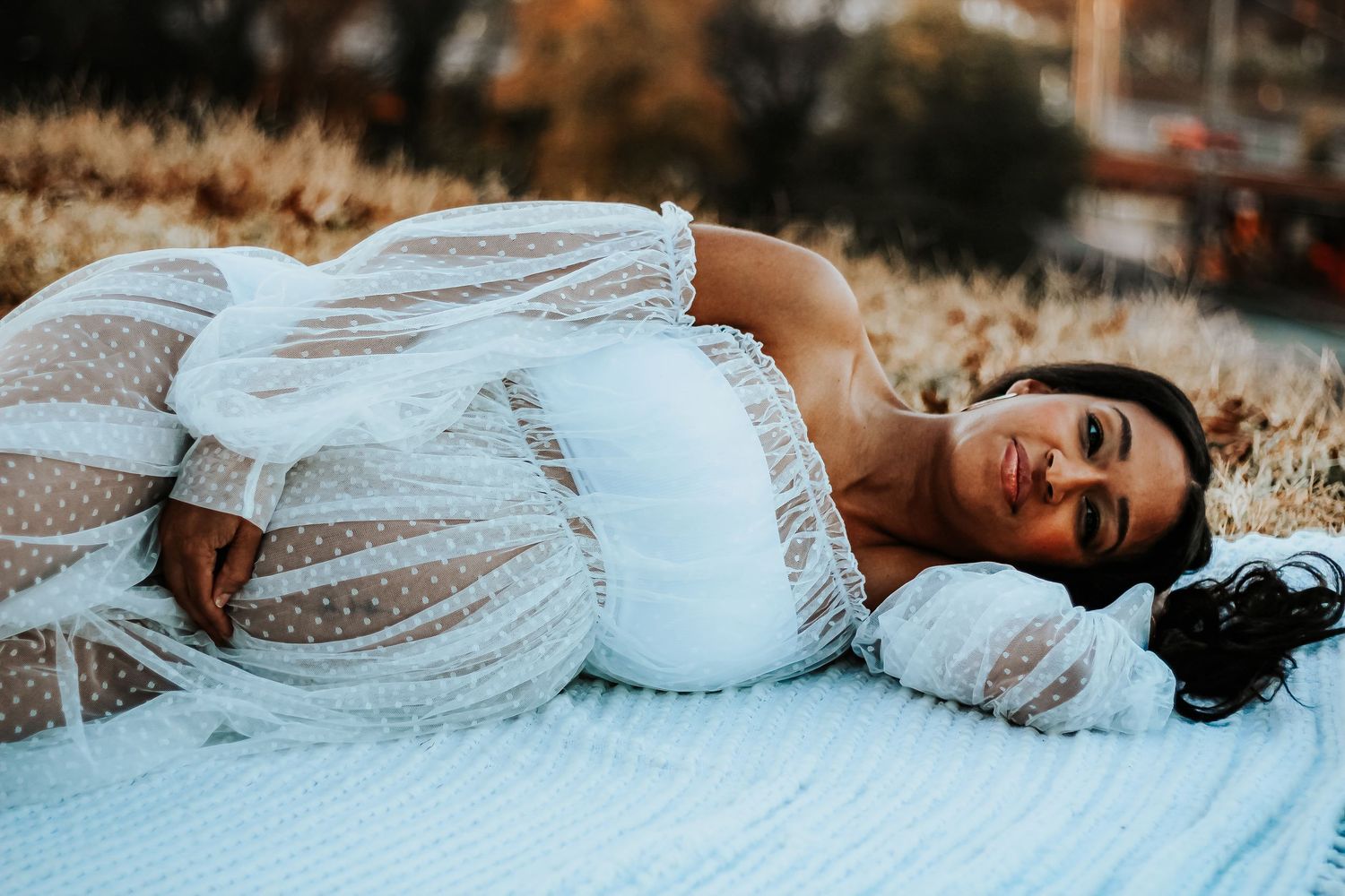 Maternity photo of woman in white lace dress lying on blanket during golden hour.