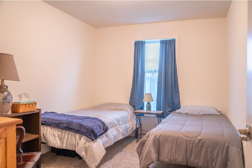 Basic bedroom with two single beds, blue curtains, and a wooden desk beside beige walls.