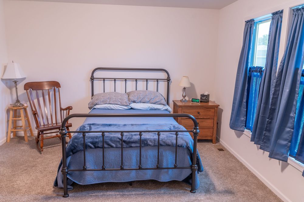 Cozy bedroom with metal frame bed, wooden nightstands, blue curtains and a rocking chair in the corner.