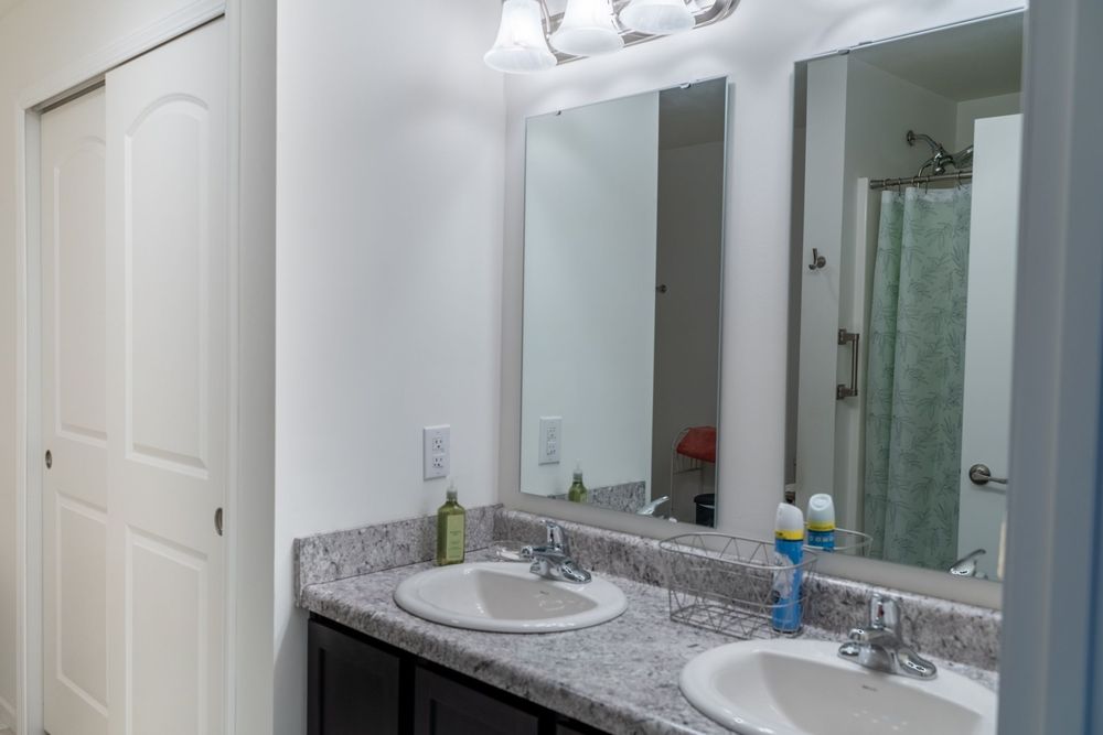 Modern bathroom vanity with dual sinks, granite countertops, large mirrors and white cabinets under bright lighting.