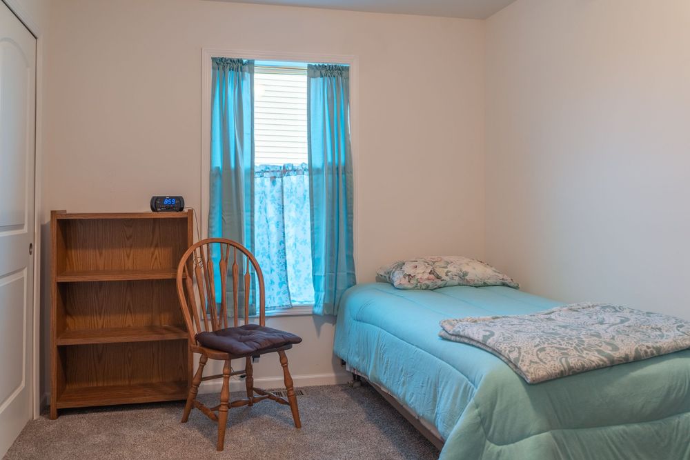 Small bedroom with twin bed, wooden bookshelf, chair and turquoise curtains in a minimalist setting.