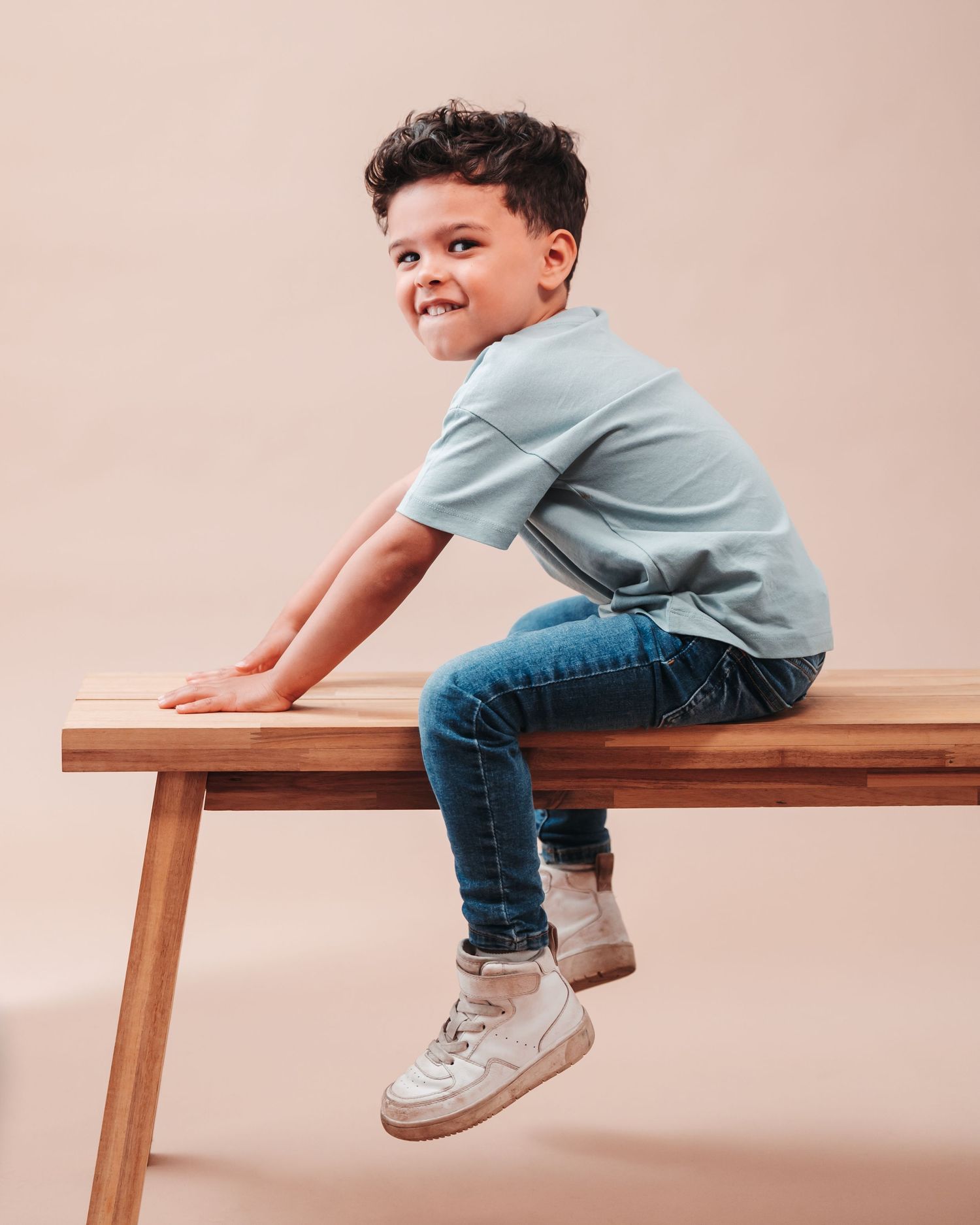 Child in light blue t-shirt and jeans sitting casually on wooden bench against pink background.