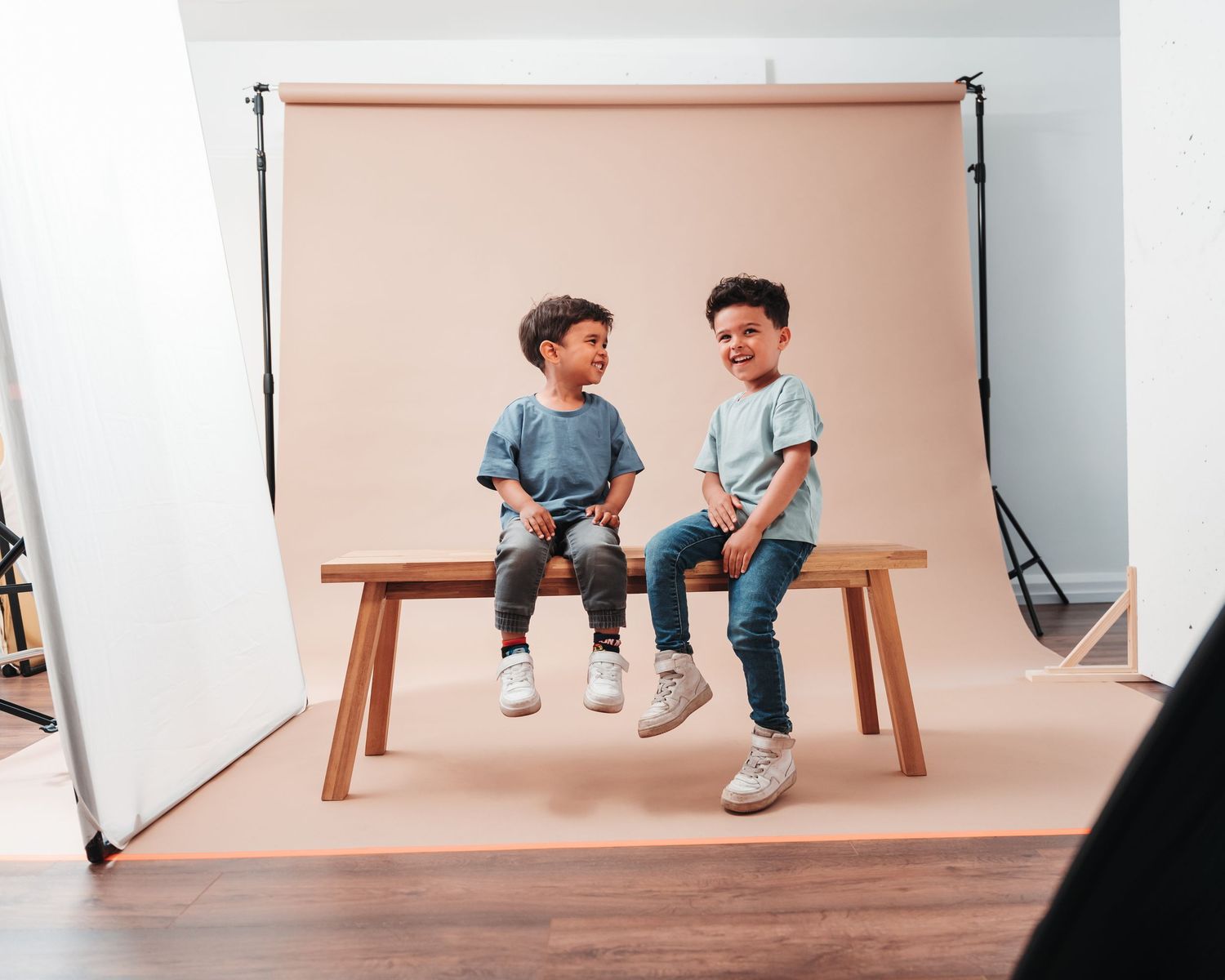 Two children sitting on wooden bench in photo studio with pink backdrop and visible lighting setup.