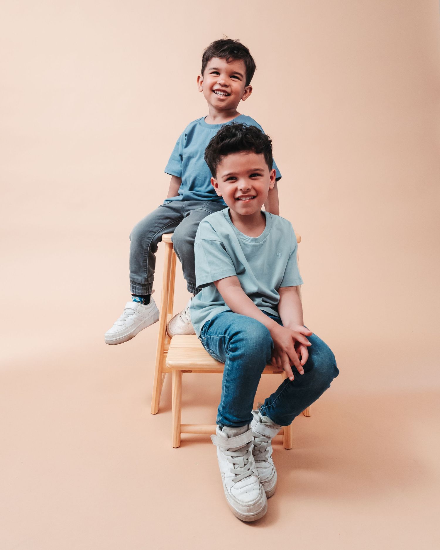 Two children in matching blue outfits posing on wooden stool against pink backdrop.