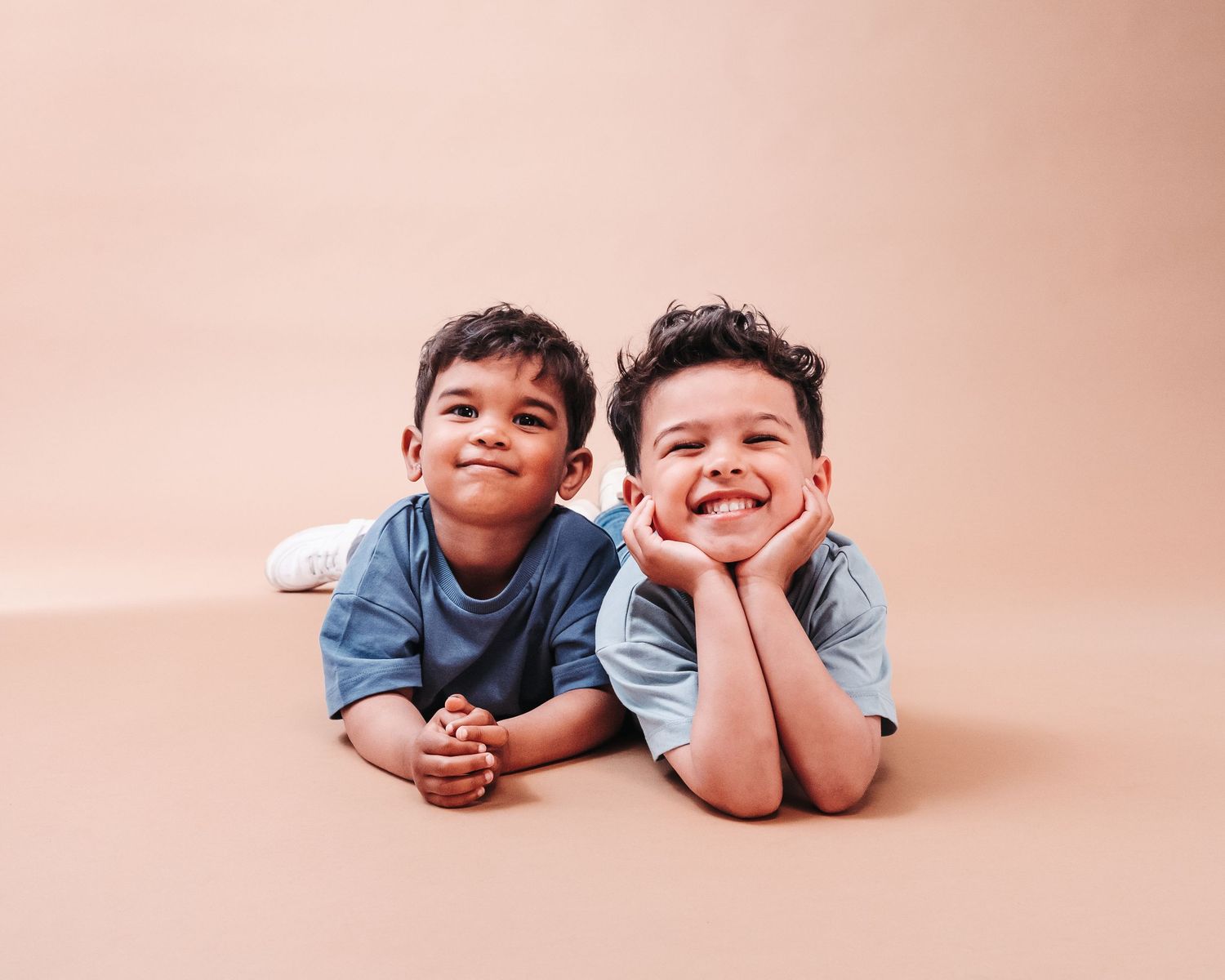 Two children in blue shirts lying on pink background showing happy expressions.