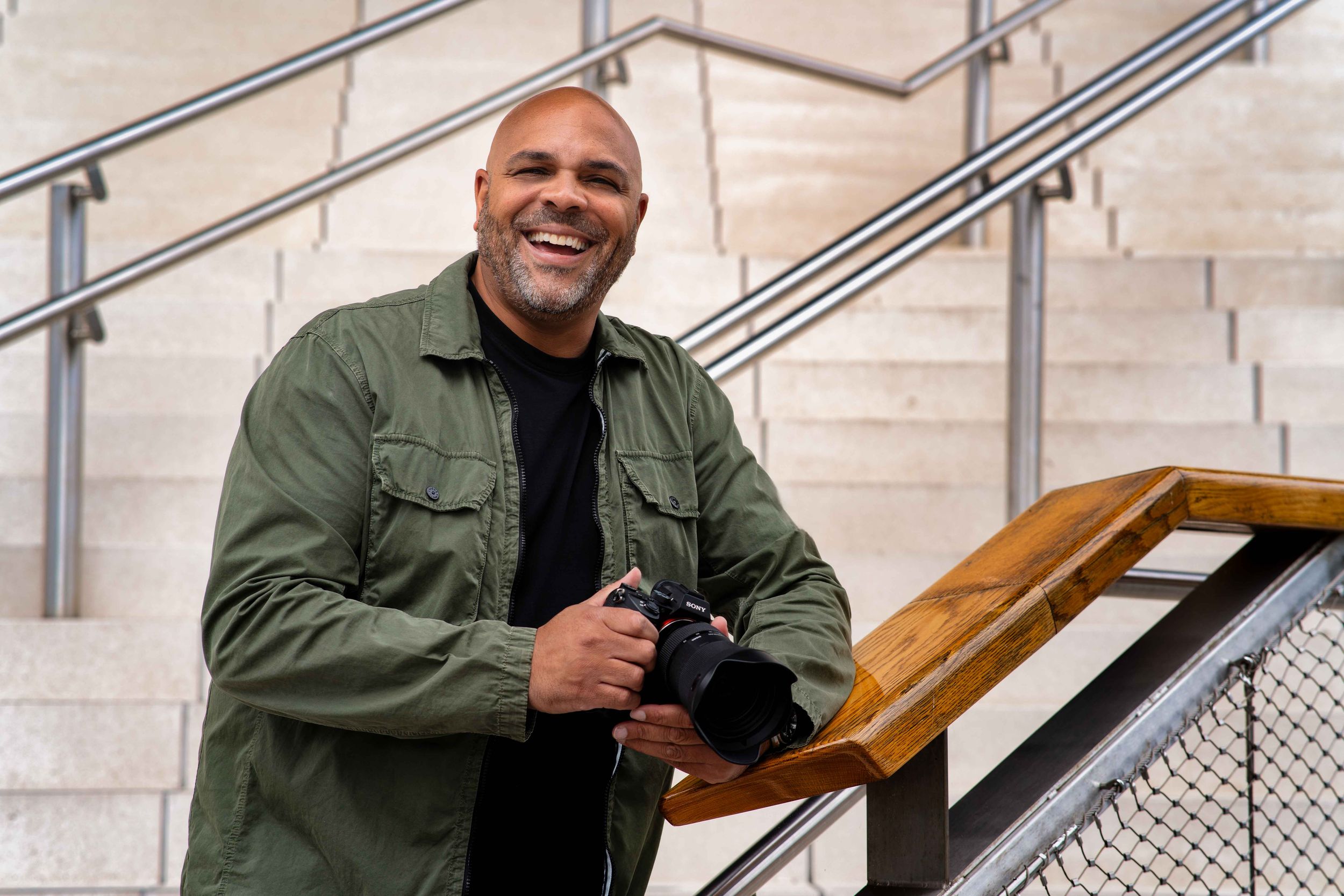 A cheerful person wearing an olive green military-style jacket leans on a wooden railing near concrete steps. He's a branding photographer