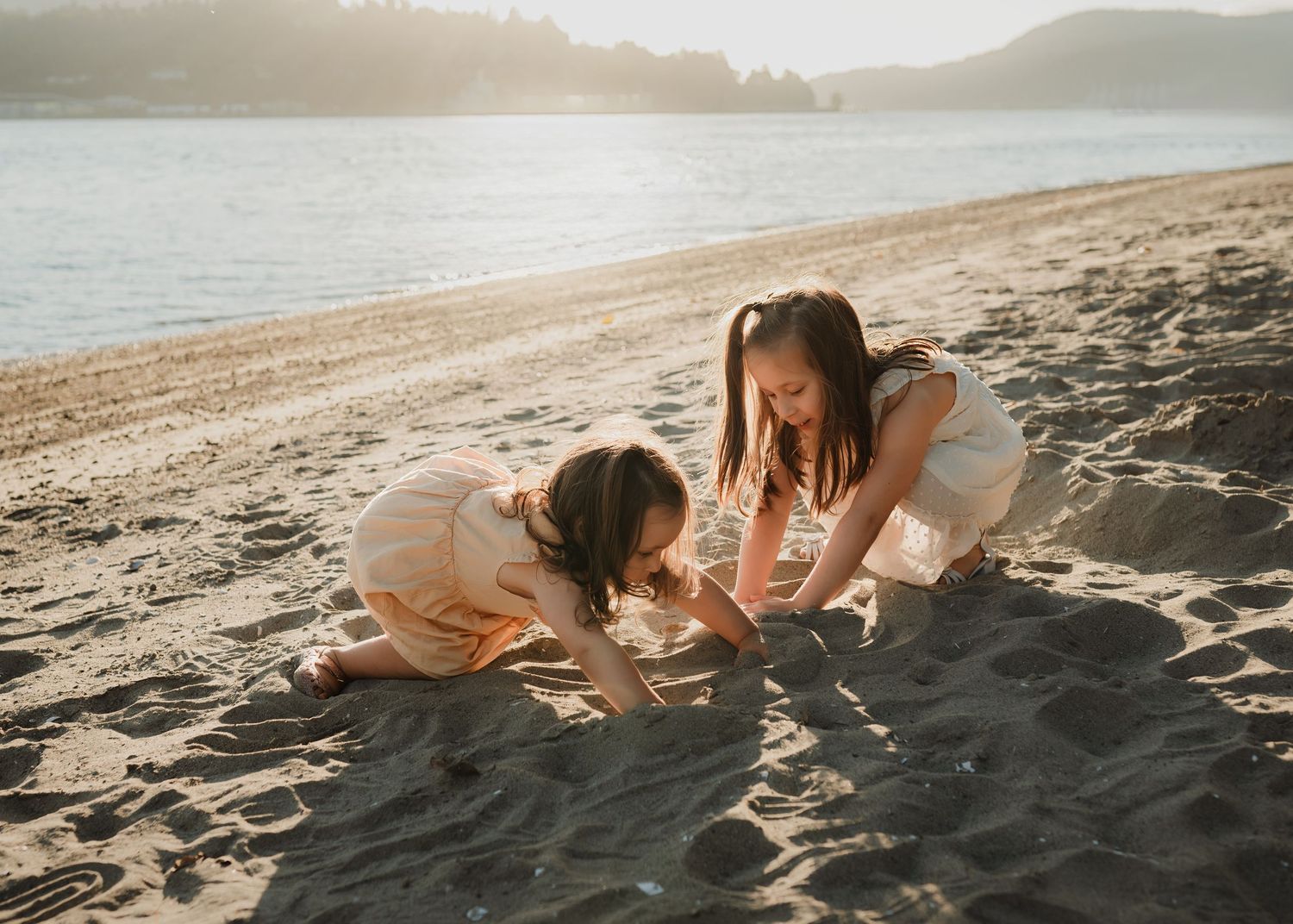Two people playing in the sand at sunset on a tranquil beach with mountains visible across the water.