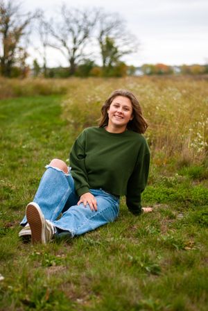 A senior girl sitting on the ground outdoors, with trees and greenery in the background