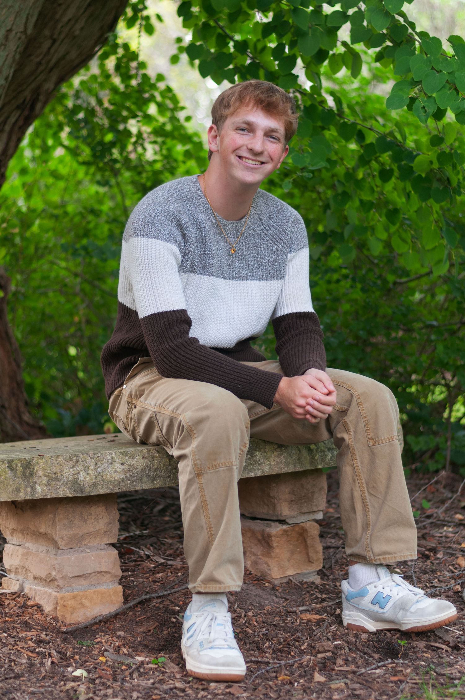 A senior boy in a sweater and khakis sitting on a bench with greenery in the background