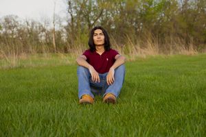 A portrait of a young man wearing a red shirt and blue jeans sitting in a field.