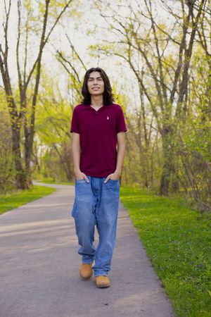 A portrait of a young man wearing a red shirt and blue jeans standing on a sidewalk with trees in the background.