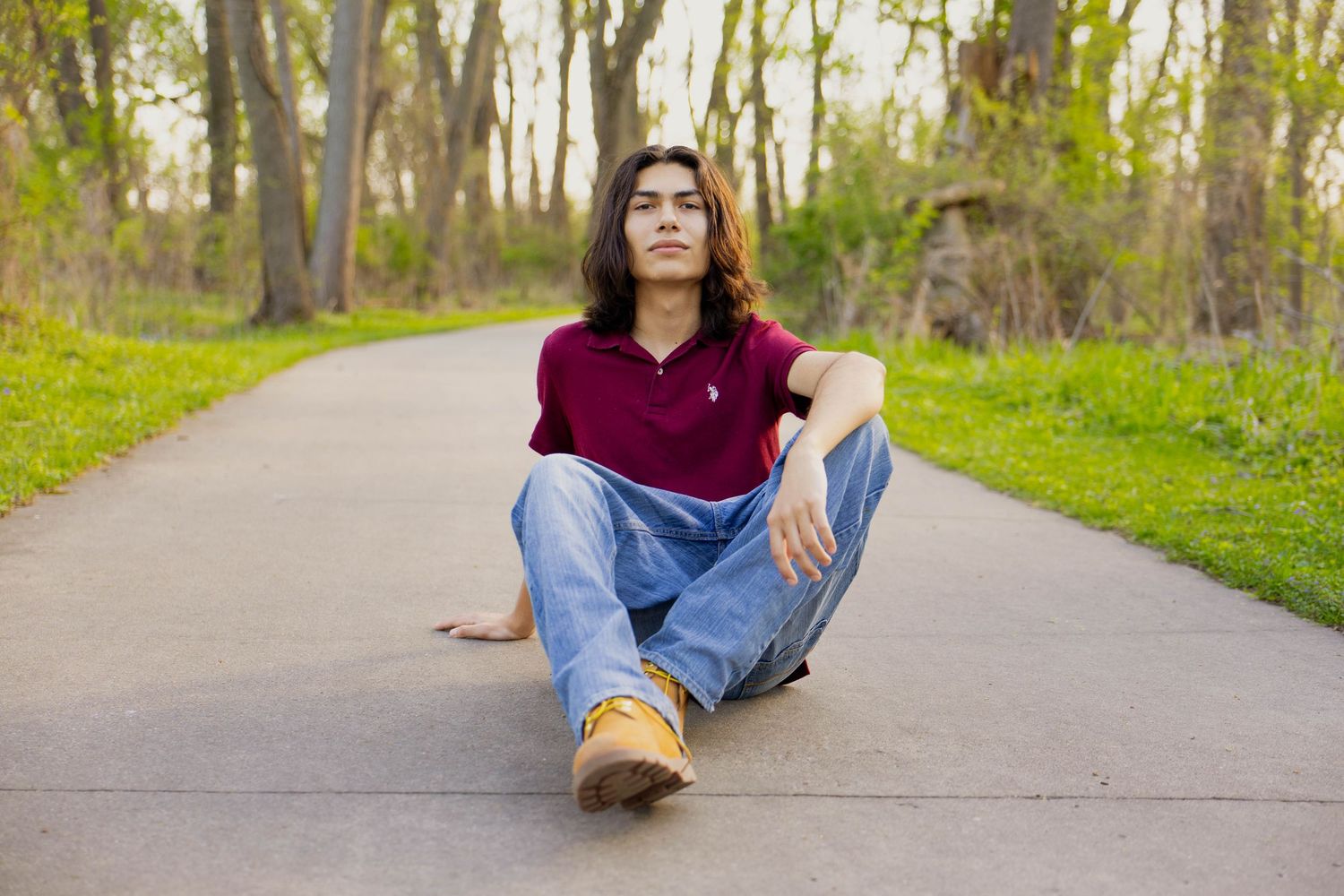 A portrait of a young man wearing a red shirt and blue jeans sitting on a sidewalk with trees in the background.