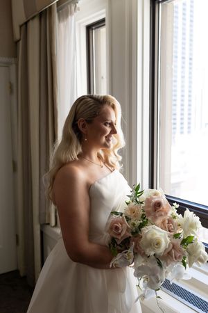 Bride in white dress holding bouquet poses by window with city views.